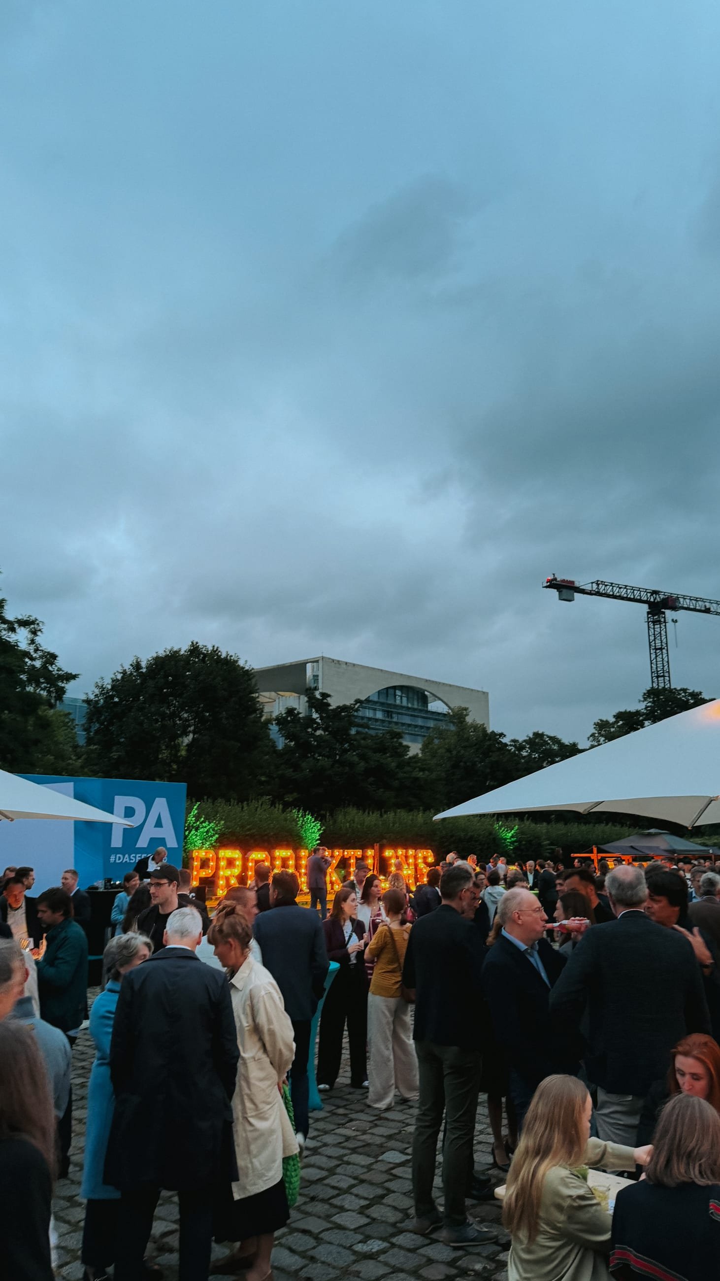 Guests in the courtyard at dusk, the illuminated „Produktionsallianz“ lettering, the Chancellery and a construction crane behind them