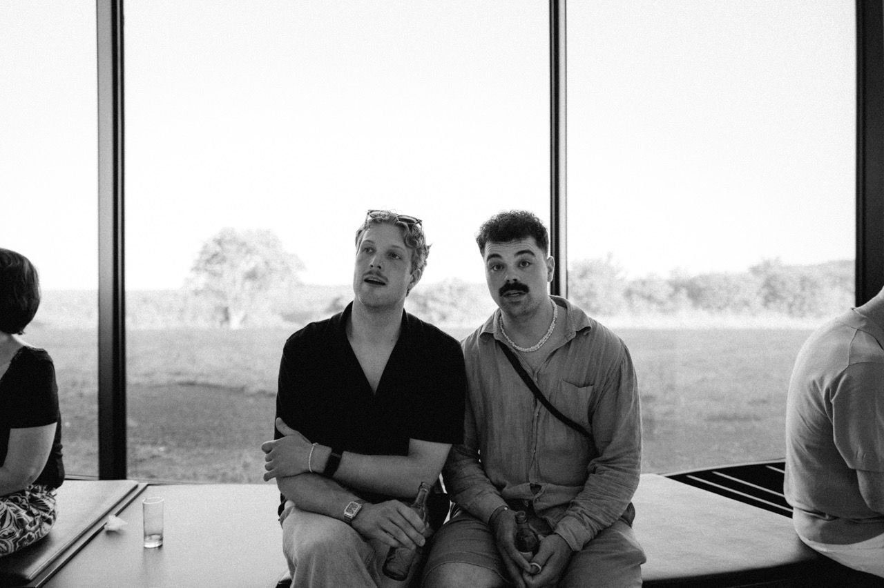 Two men sitting in front of a glass wall with open countryside behind it