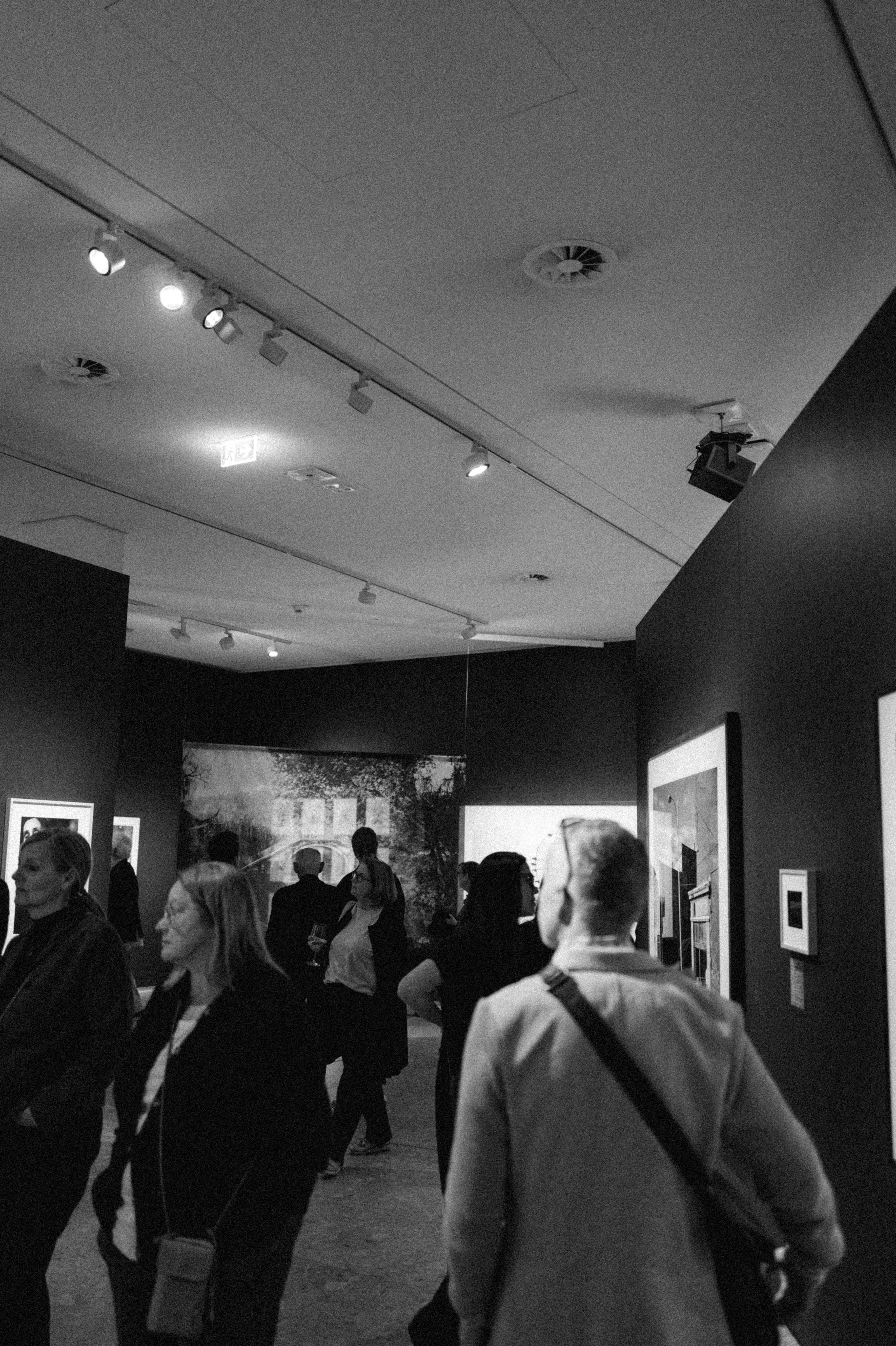 Blick in die abgedunkelten Ausstellungsräume mit Besucherinnen und Besuchern, schwarzweiß