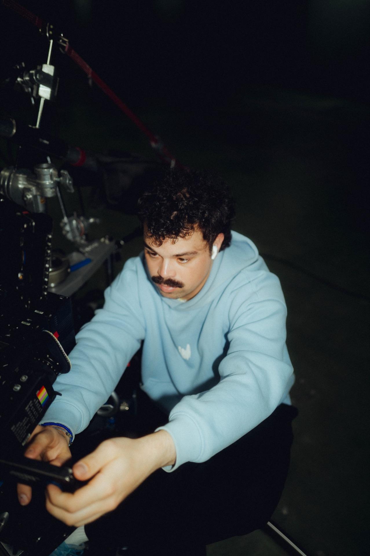 Cinematographer in a light-blue hoodie adjusts the camera with focus