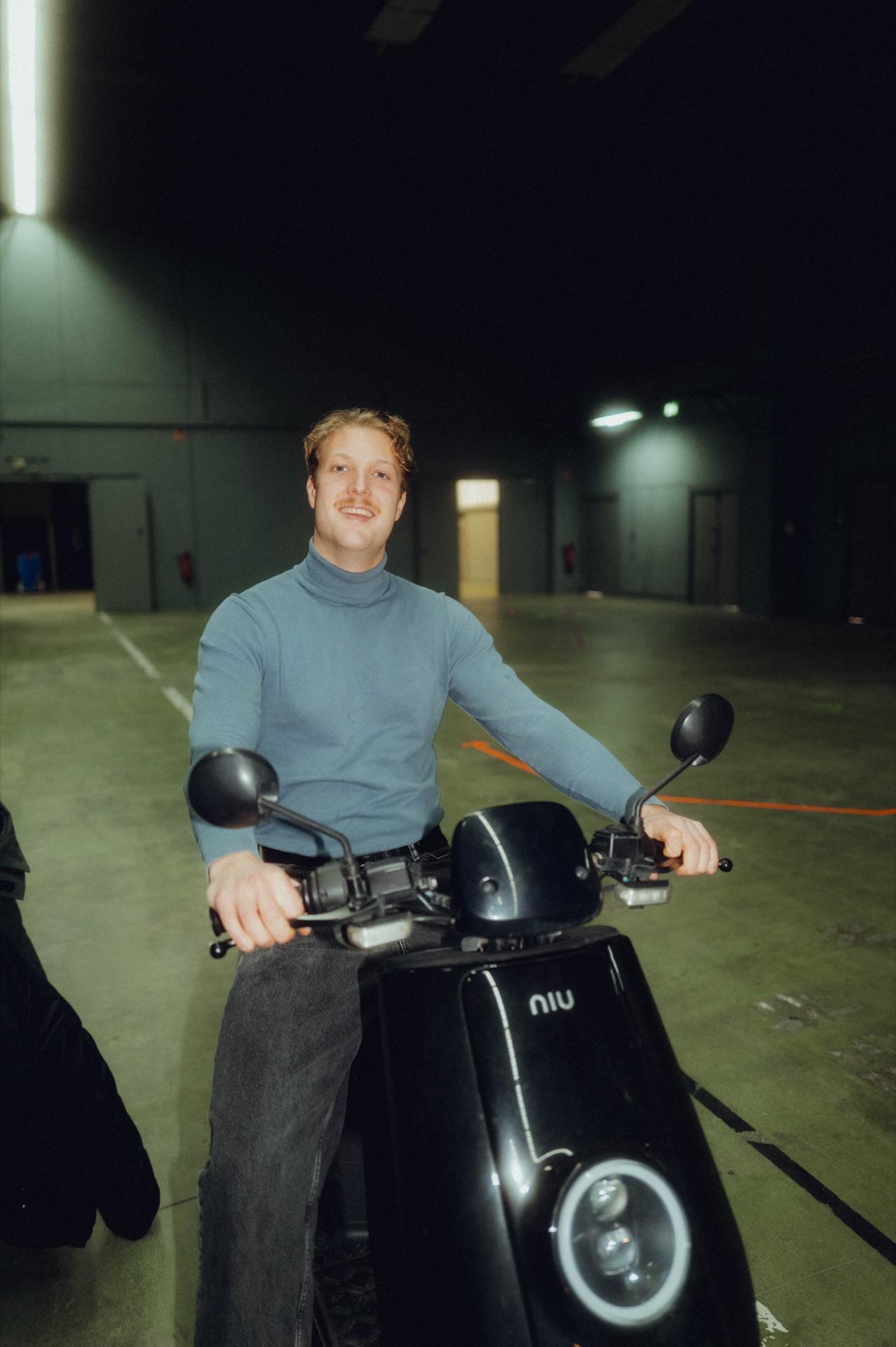 Crew member in a turtleneck sits laughing on a motor scooter in the hall