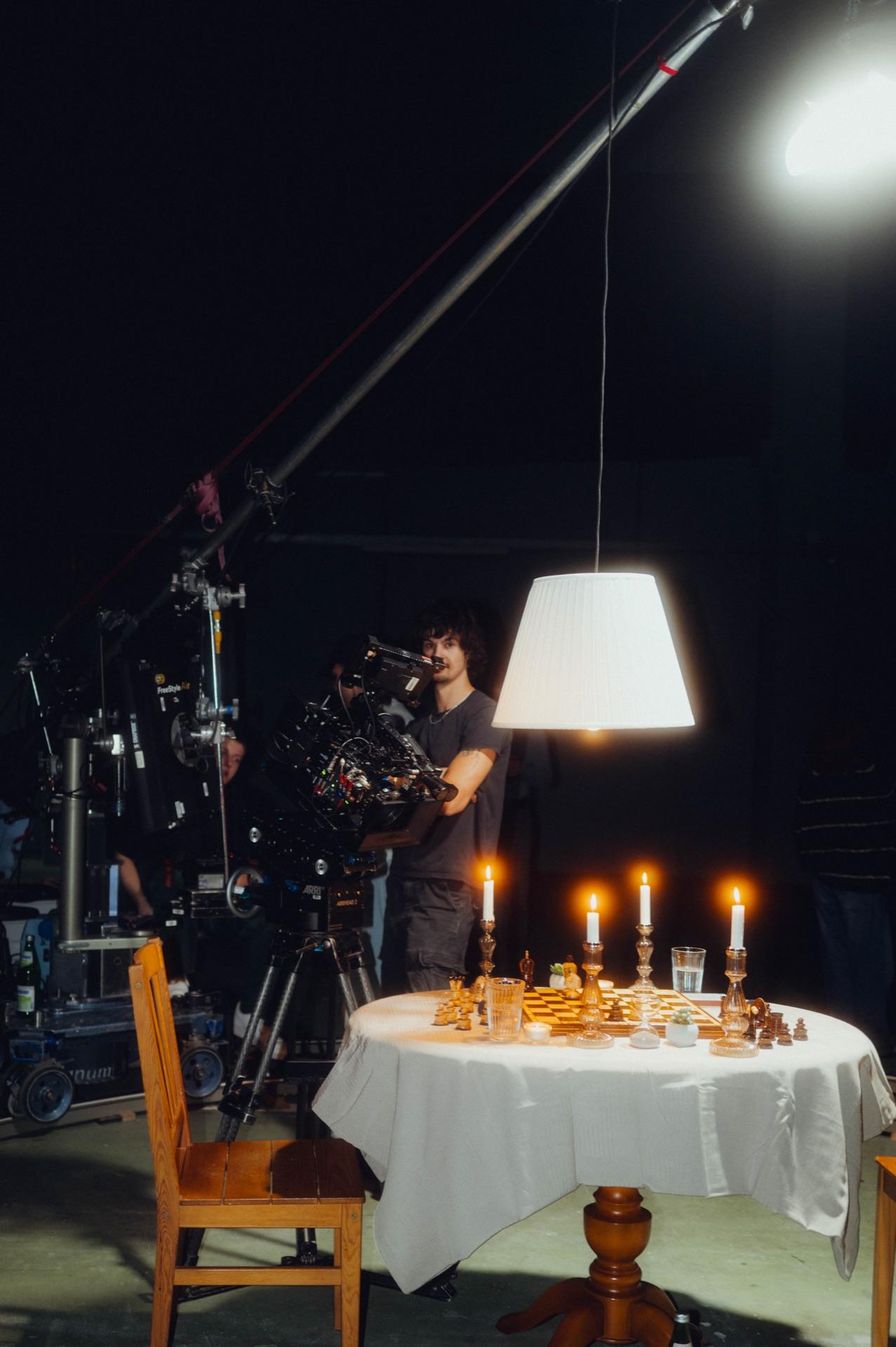 The set: the laid table with chessboard and candles under a hanging lamp, camera in front