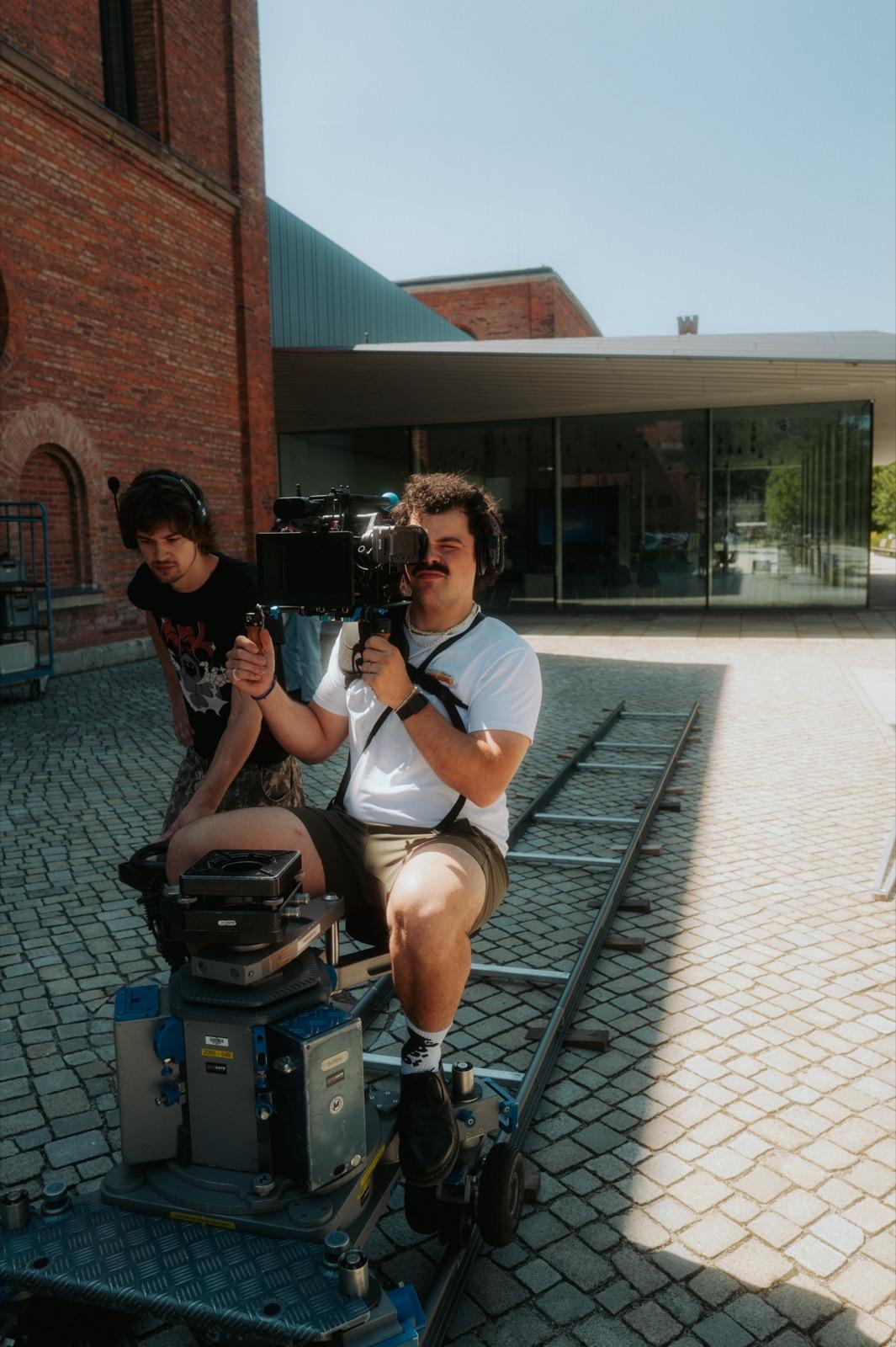 Behind the Scenes: Operator und Assistenz sitzen auf dem Dolly auf den Schienen vor einer Backsteinfassade in der Sonne