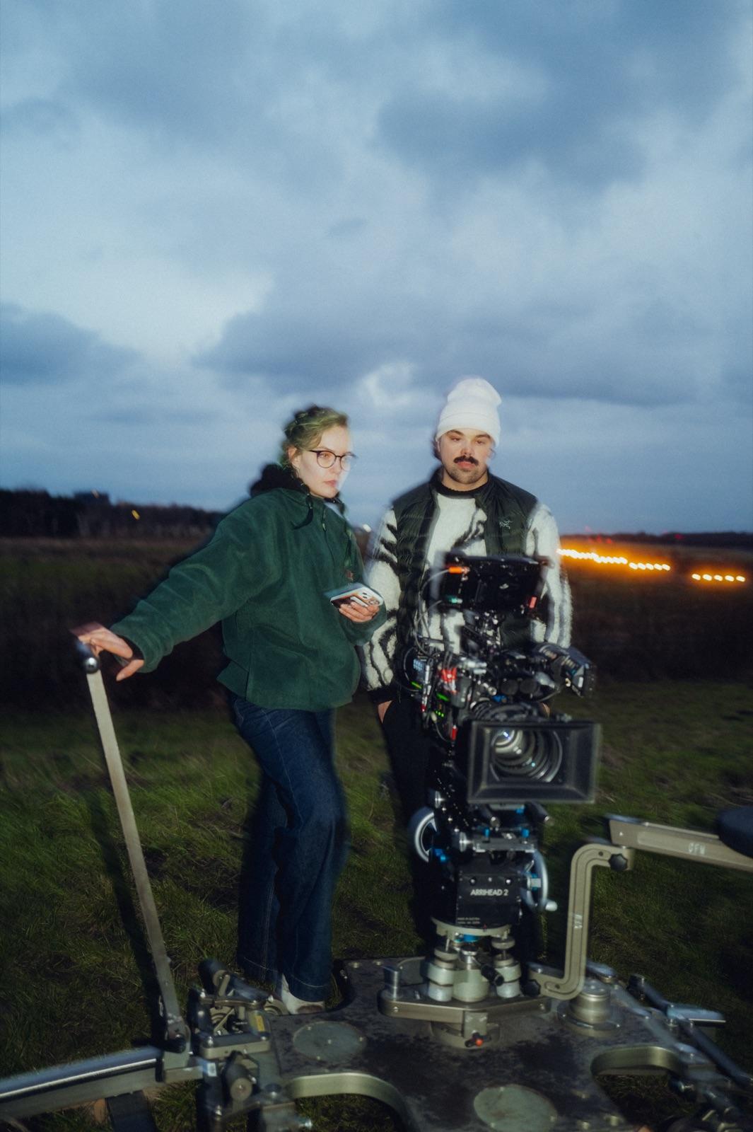Behind the Scenes: zwei Crew-Mitglieder auf der Dolly-Plattform blicken zur Kamera, Runway-Lichter dahinter