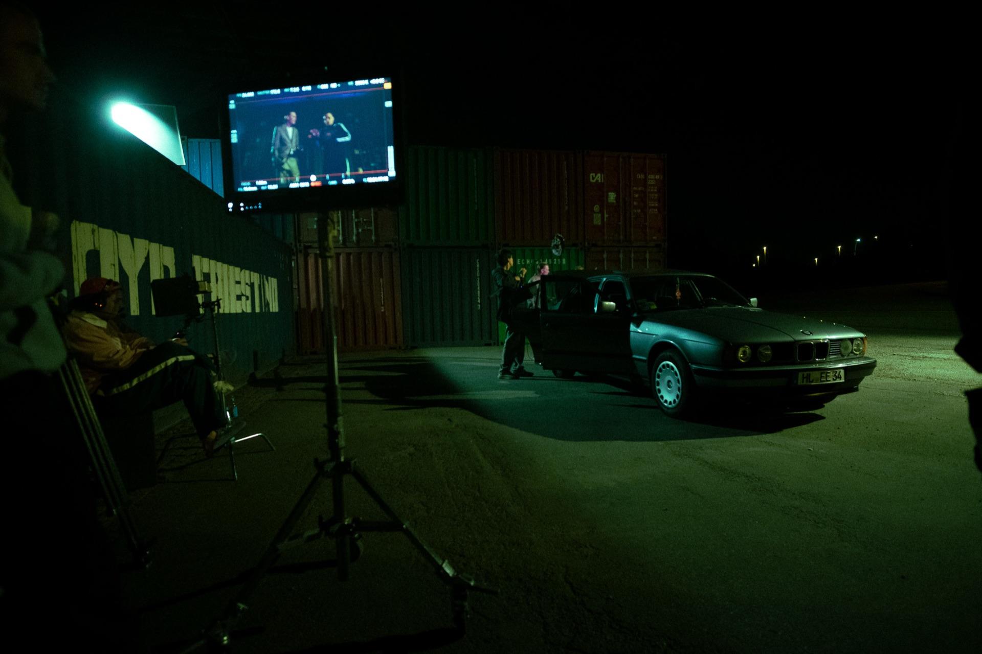 Behind the scenes: set overview with a monitor on the stand and the crew at the old BMW in front of the containers