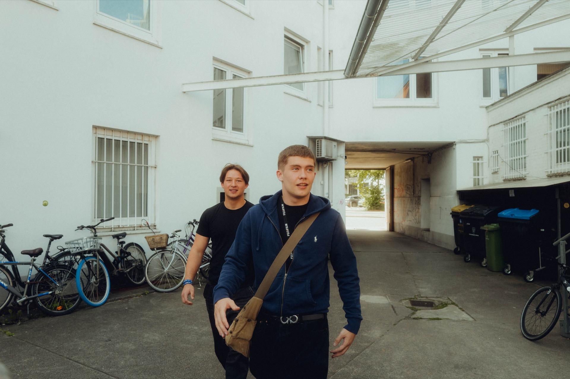 Behind the scenes: two young men walk through a Hamburg backyard, a row of parked bikes on the left, a gateway in the background