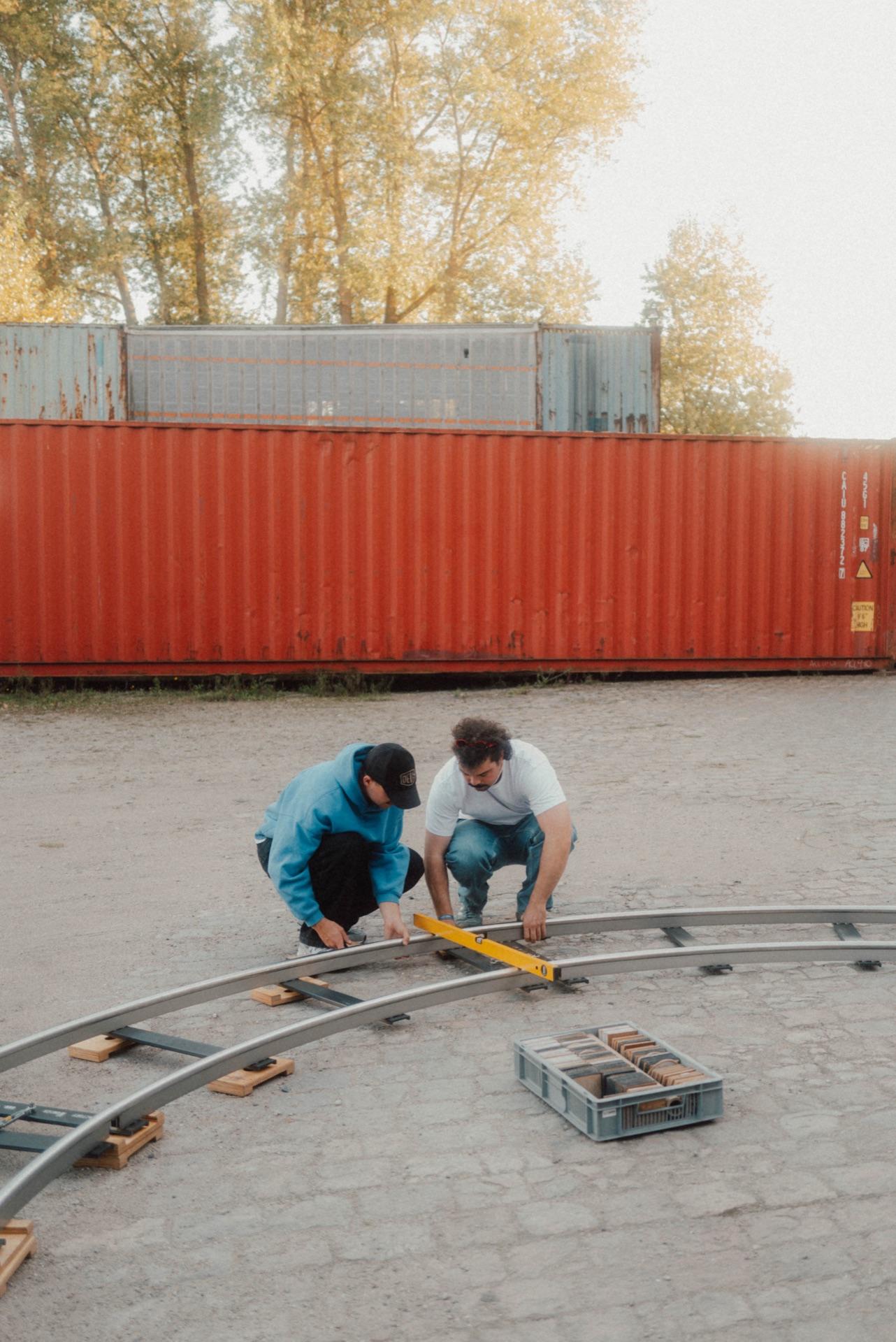 Zwei Crew-Mitglieder verlegen die gebogenen Dolly-Schienen vor einem orangefarbenen Container, Herbstbäume in der Dämmerung
