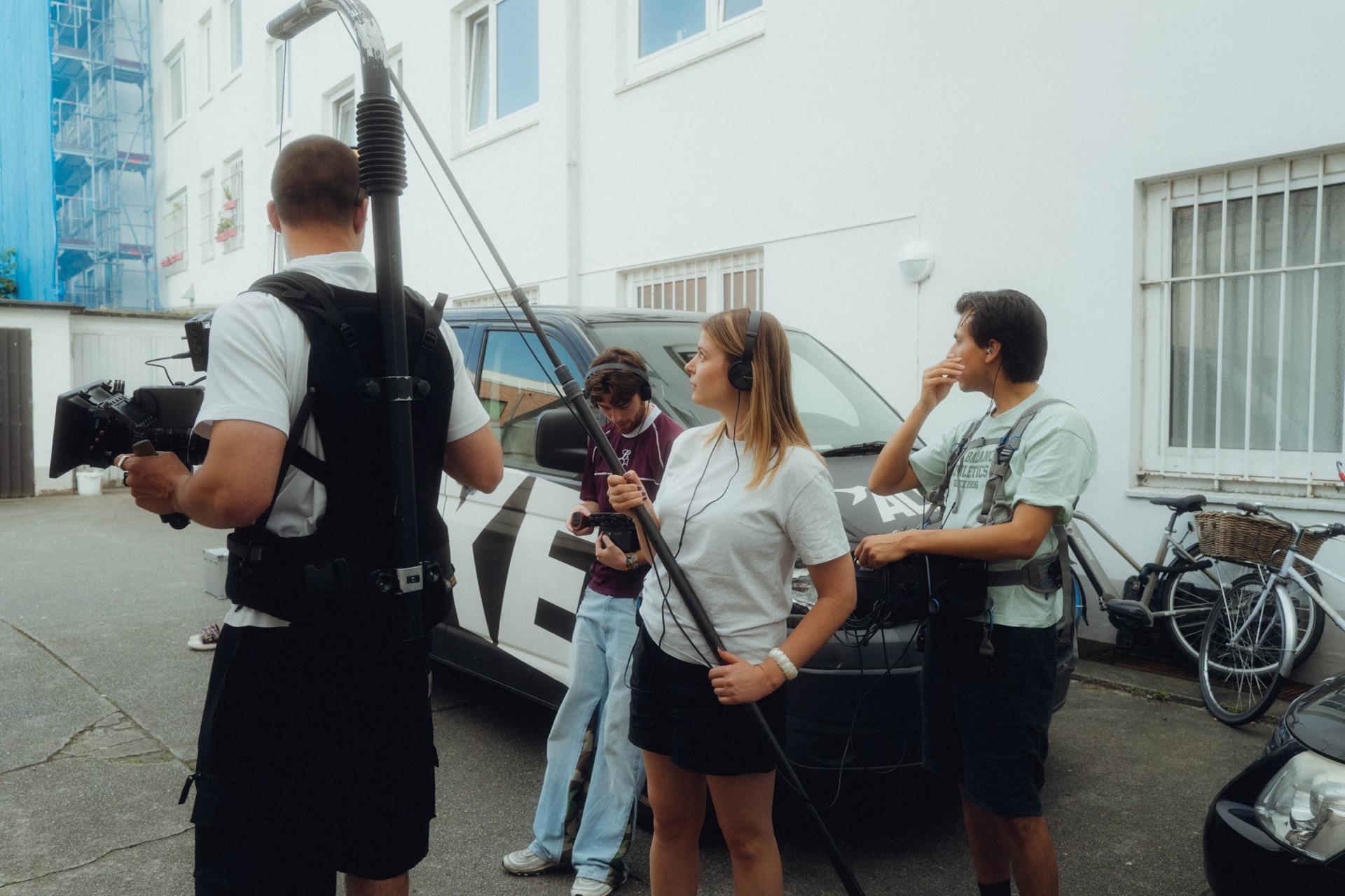 Behind the scenes: a camera operator from behind next to a boom pole, the crew filming a scene at a silver car beyond