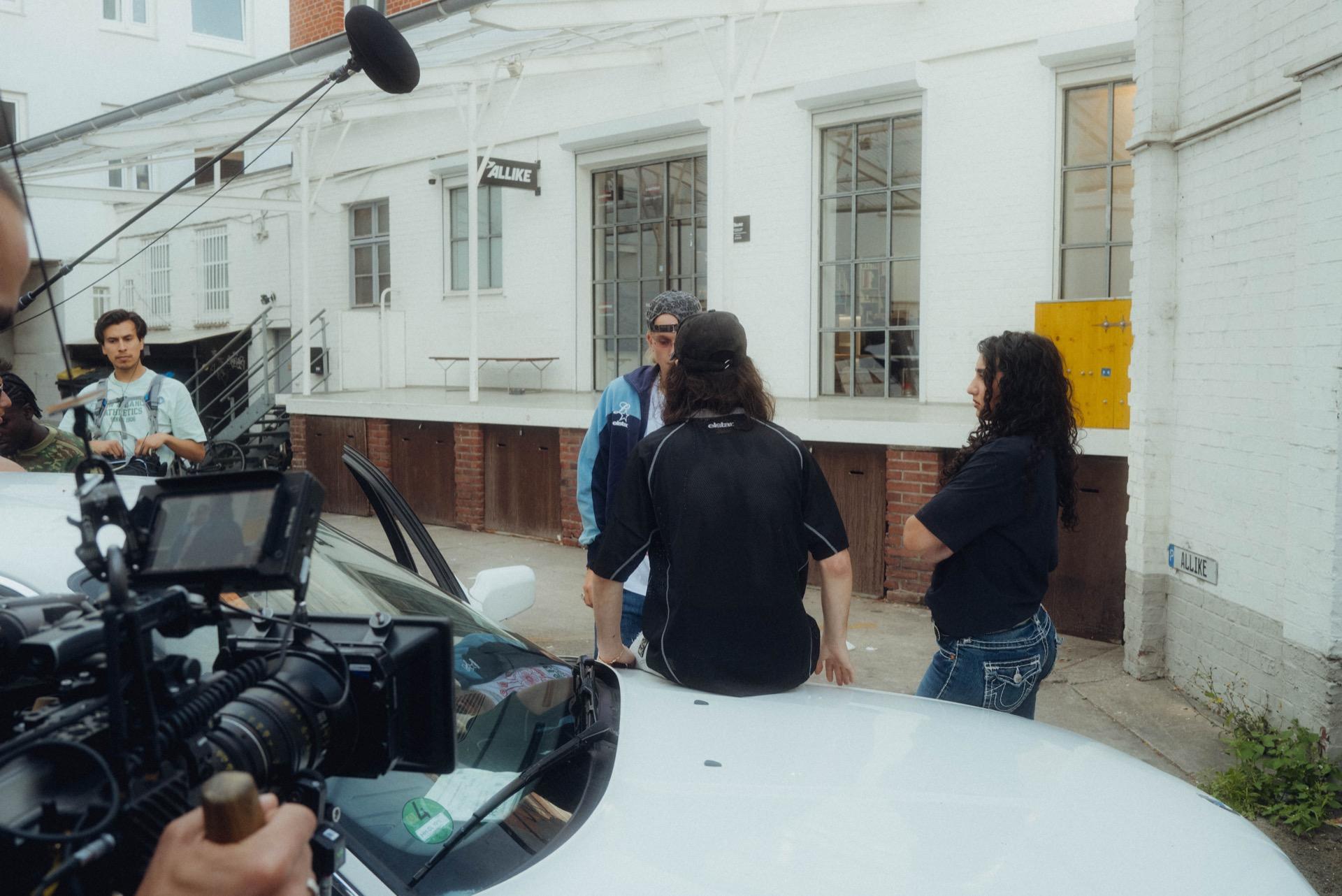 Behind the scenes: a film camera on a rig in the foreground, the crew shooting in front of a white factory facade reading ALIKE