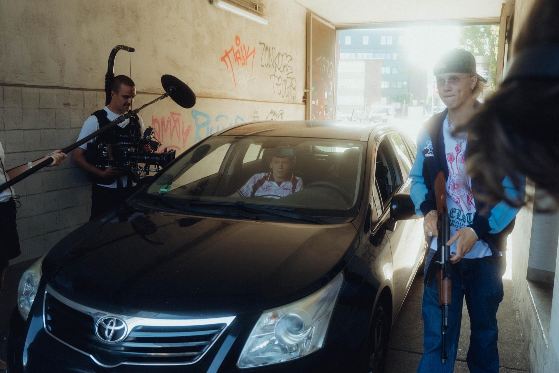 Behind the scenes: in a backlit gateway the crew films the black Toyota, a young man in a light jacket holding a prop on the right