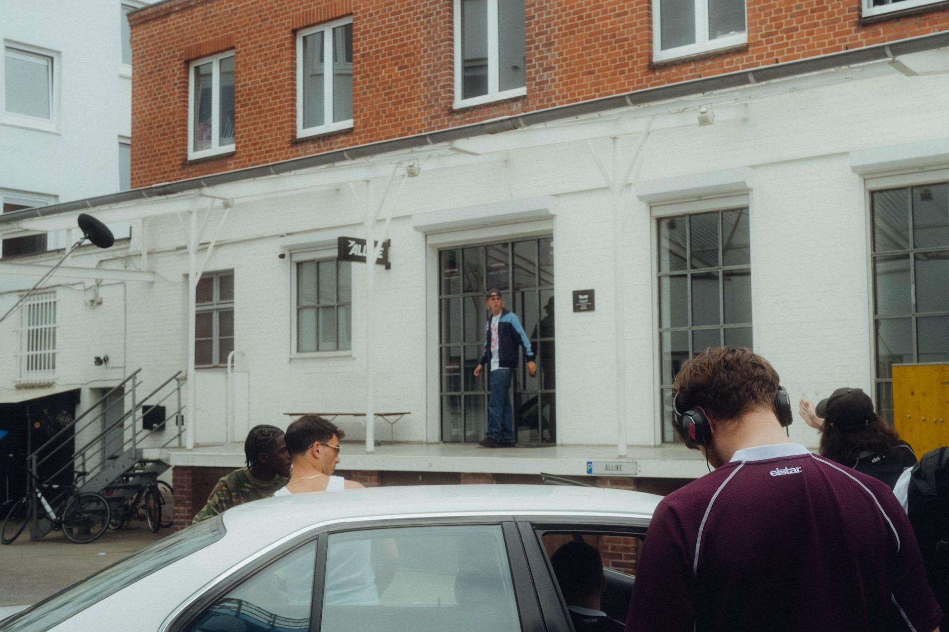 Behind the scenes: a wide view of the set, an actor standing in the glass entrance of the white hall, the crew and a white car in front