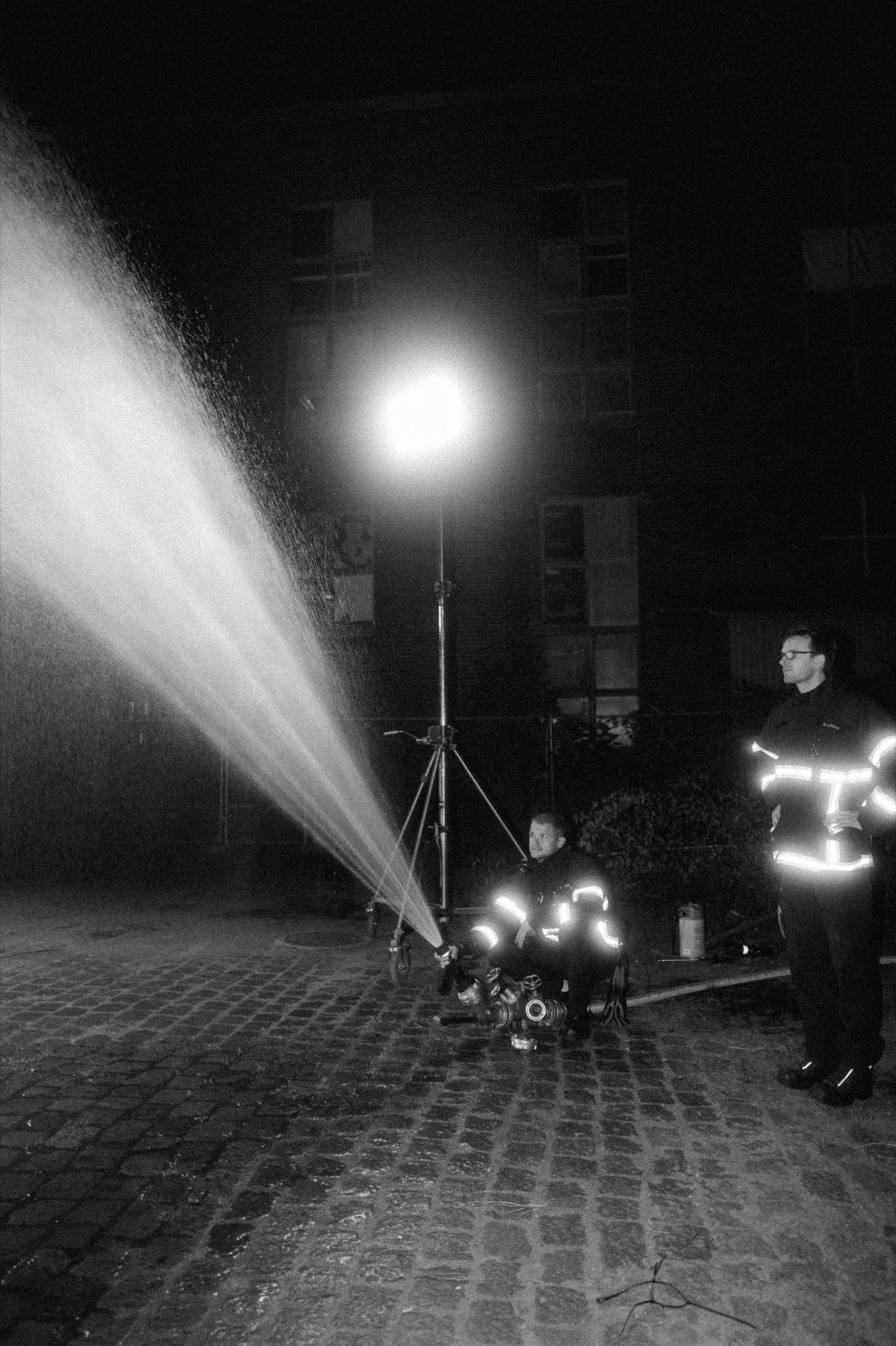Behind the Scenes: ein Scheinwerfer auf Stativ, davor sprüht ein Wasserstrahl den künstlichen Regen schräg durch die Nacht