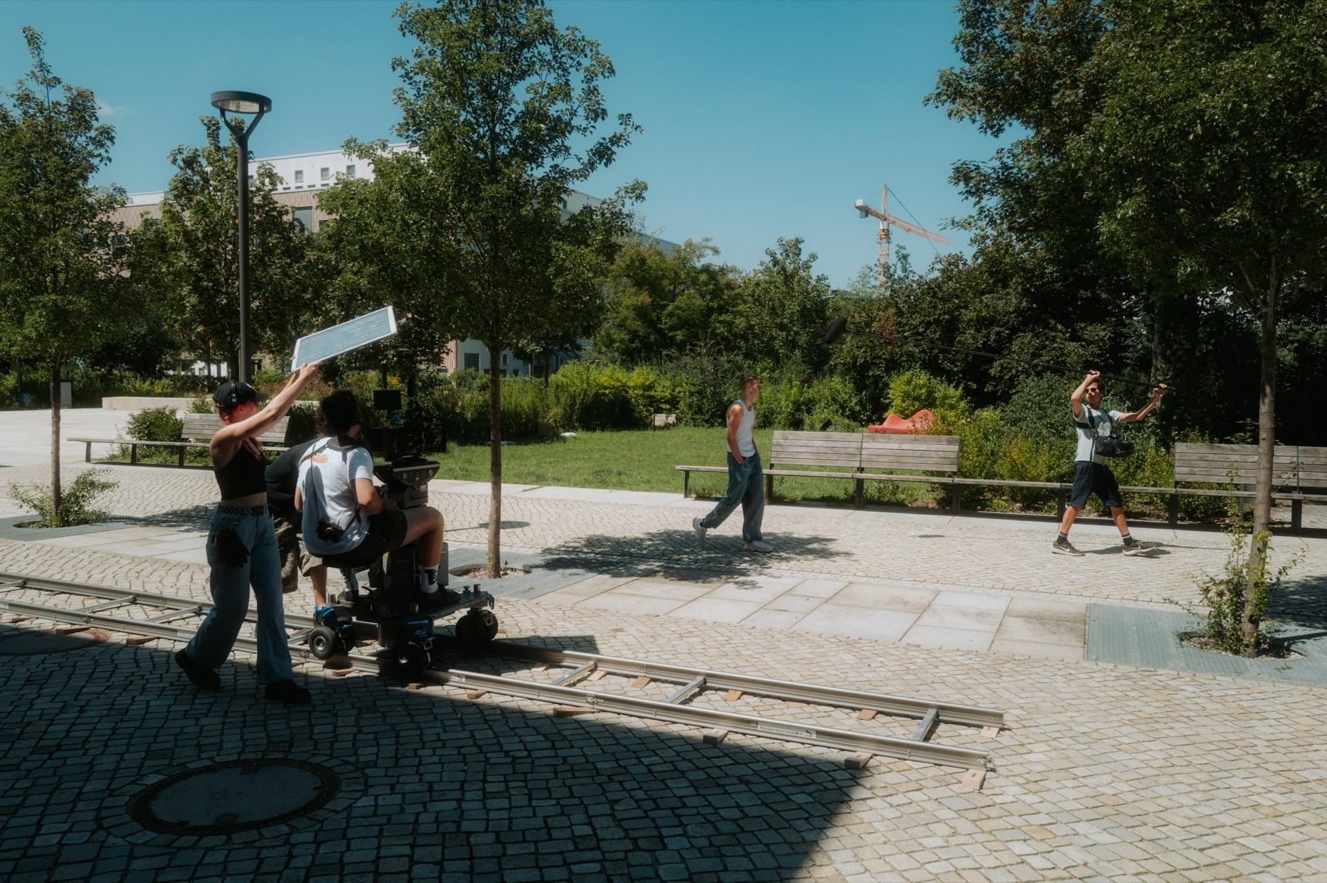 Behind the Scenes: weite Set-Übersicht im sonnigen Park, das Team an Dolly und Kamera auf den Schienen, ein Crew-Mitglied hält einen Reflektor, im Hintergrund spielt der Darsteller