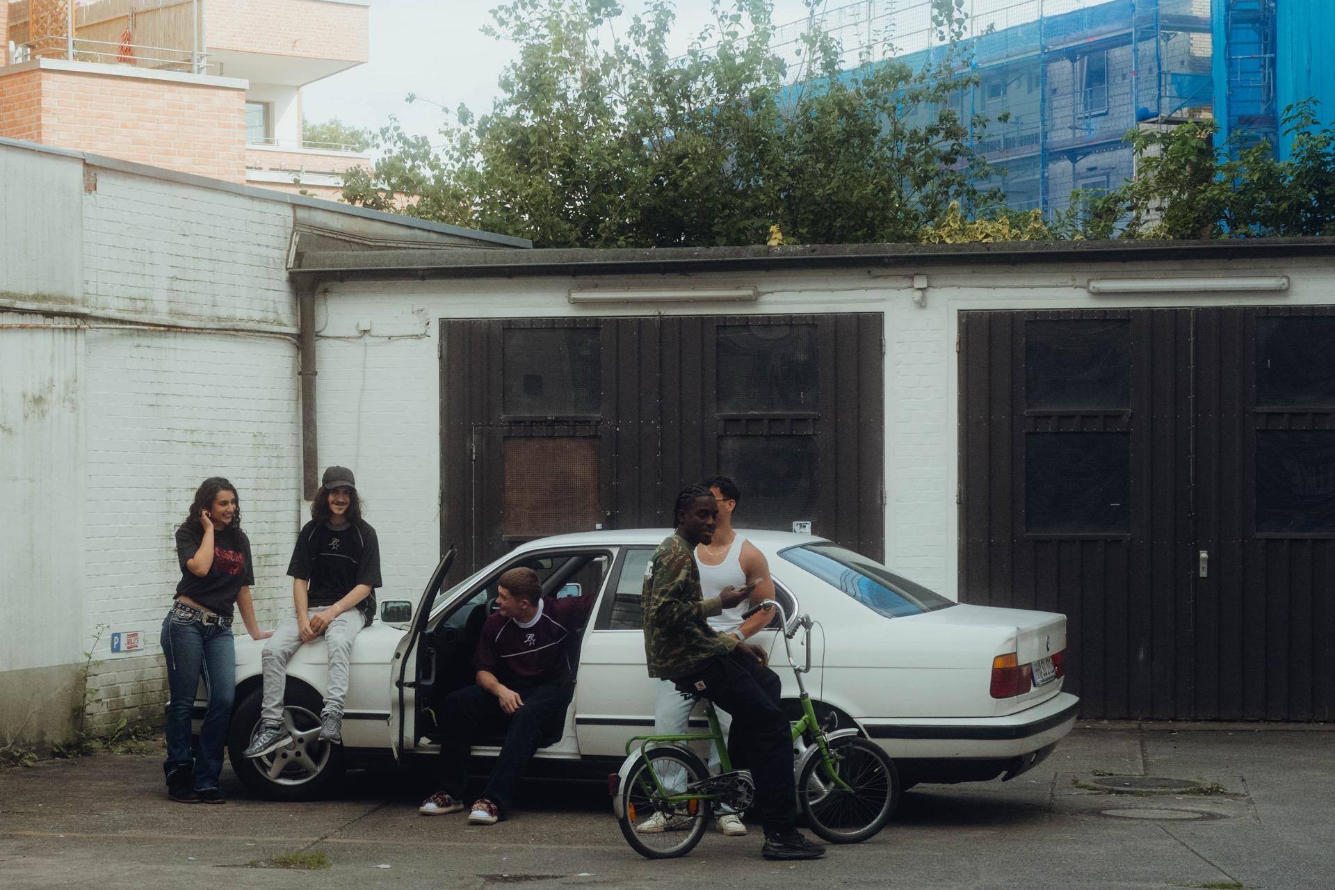 Behind the scenes: several cast members lean against an old white BMW in front of garage doors, one sitting on a folding BMX bike