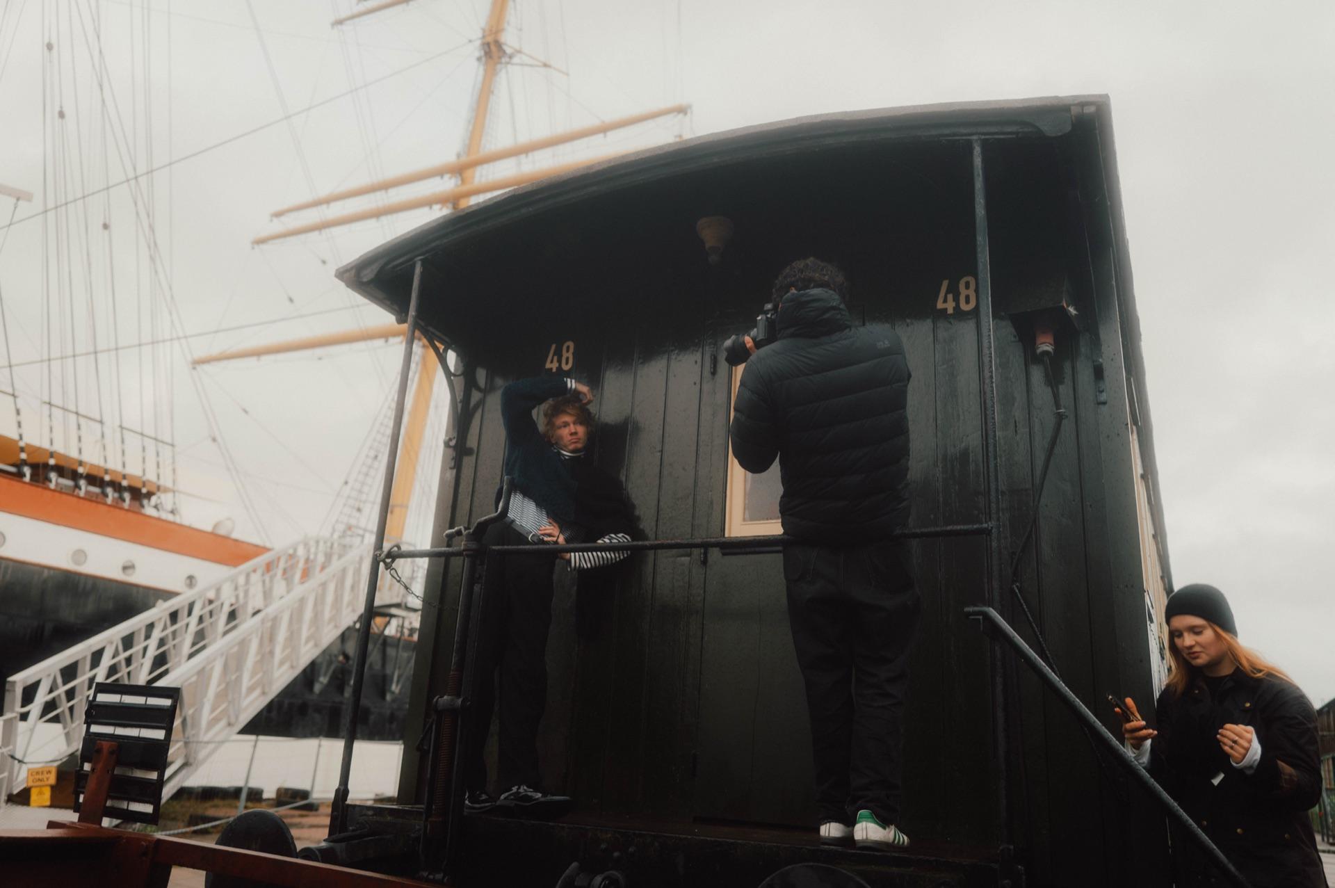 Behind the Scenes: der Fotograf richtet die Kamera auf Filip Schnack an Deck eines Schiffs, eine Person beobachtet die Szene