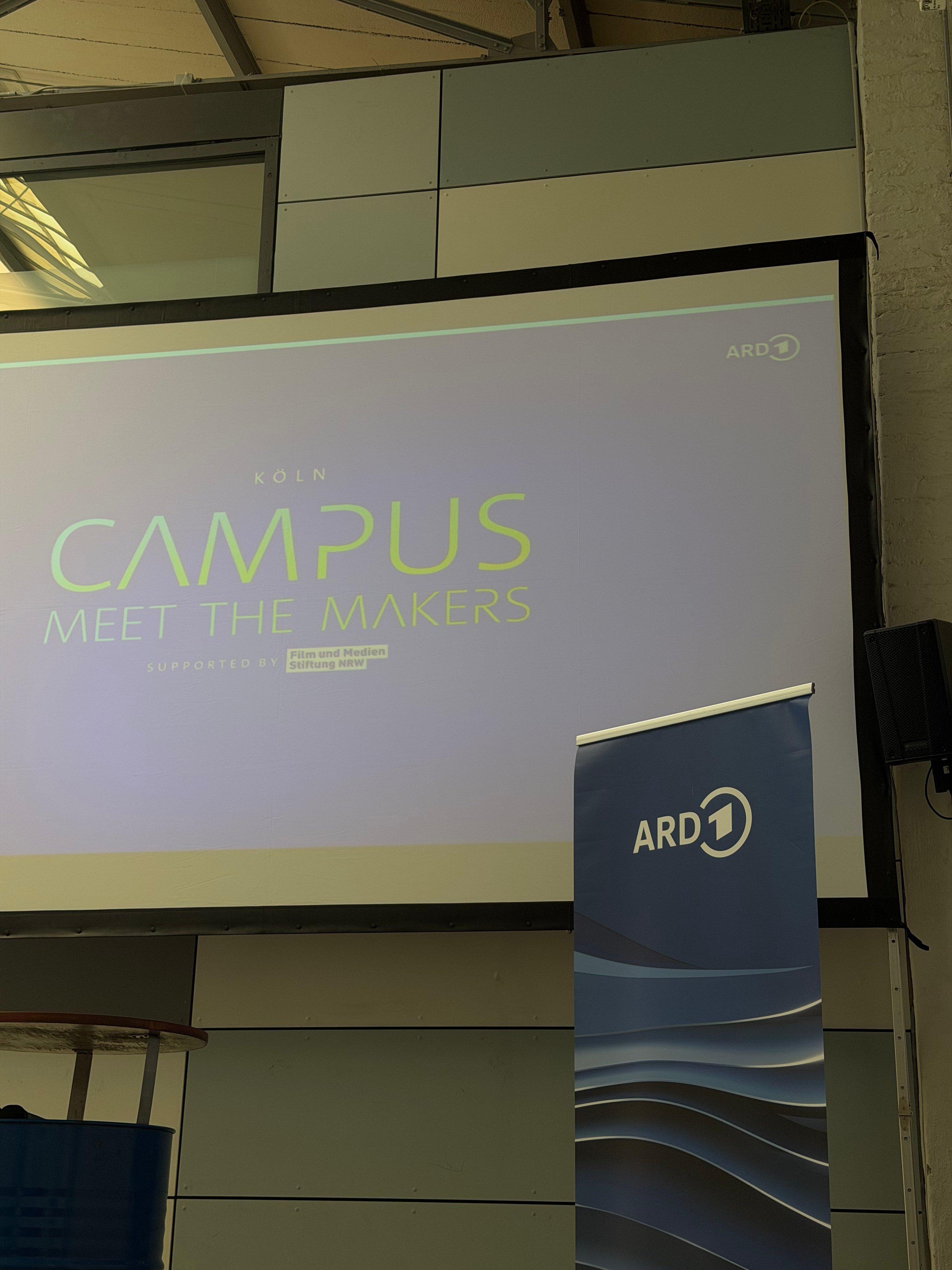 Projizierte Titelfolie „Köln Campus, Meet the Makers“ auf einer großen Leinwand, daneben ein ARD-Banner