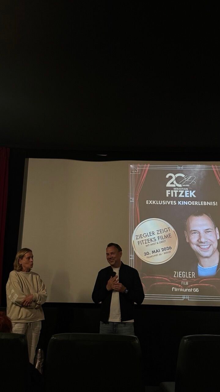 Zwei Personen stehen vor der Kinoleinwand, auf der das Plakat „20 Jahre Sebastian Fitzek, Ziegler zeigt Fitzeks Filme“ zu sehen ist