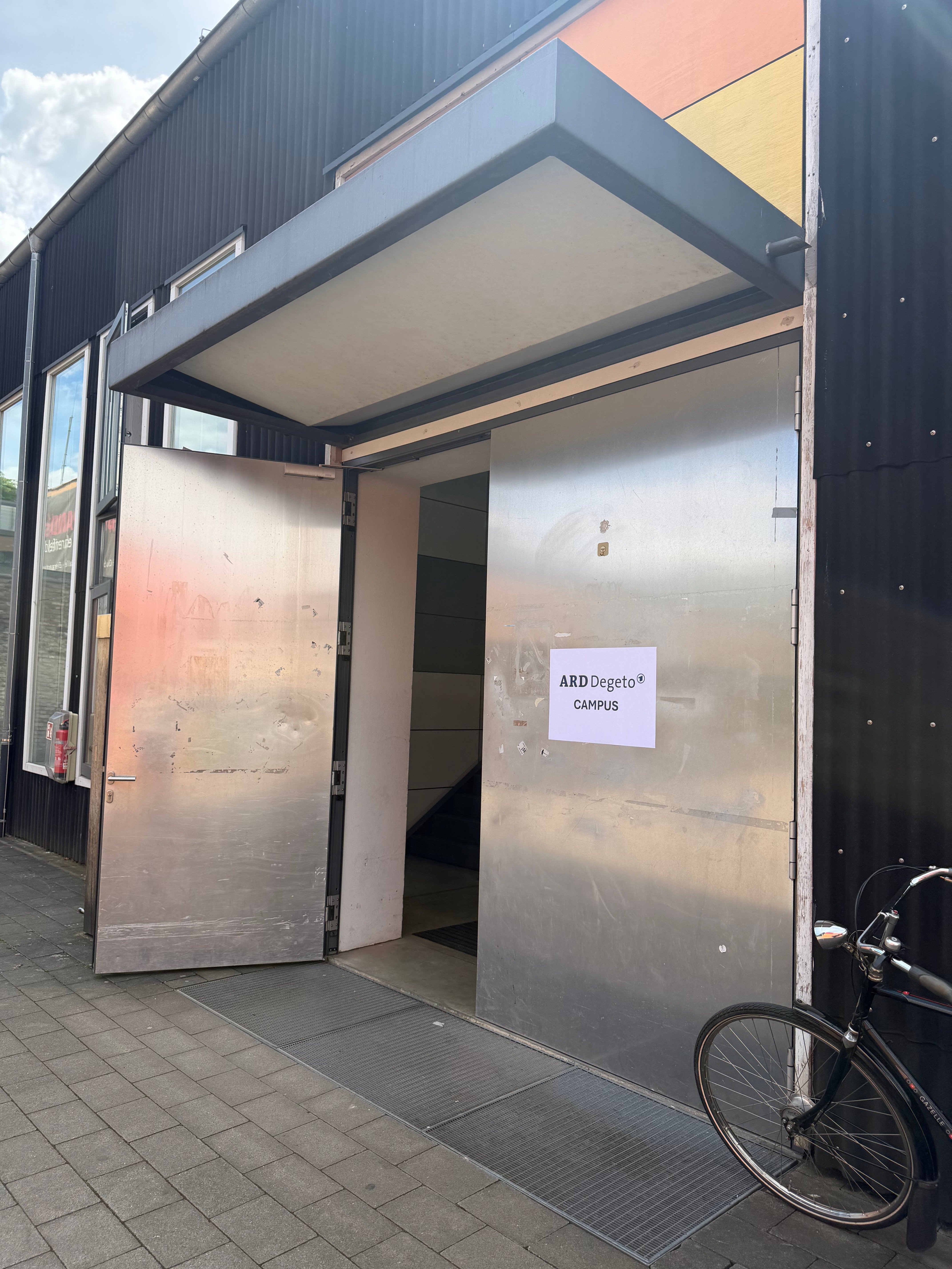 Geöffnete Metalltür mit einem Schild „ARD Degeto Campus“, davor ein angelehntes Fahrrad