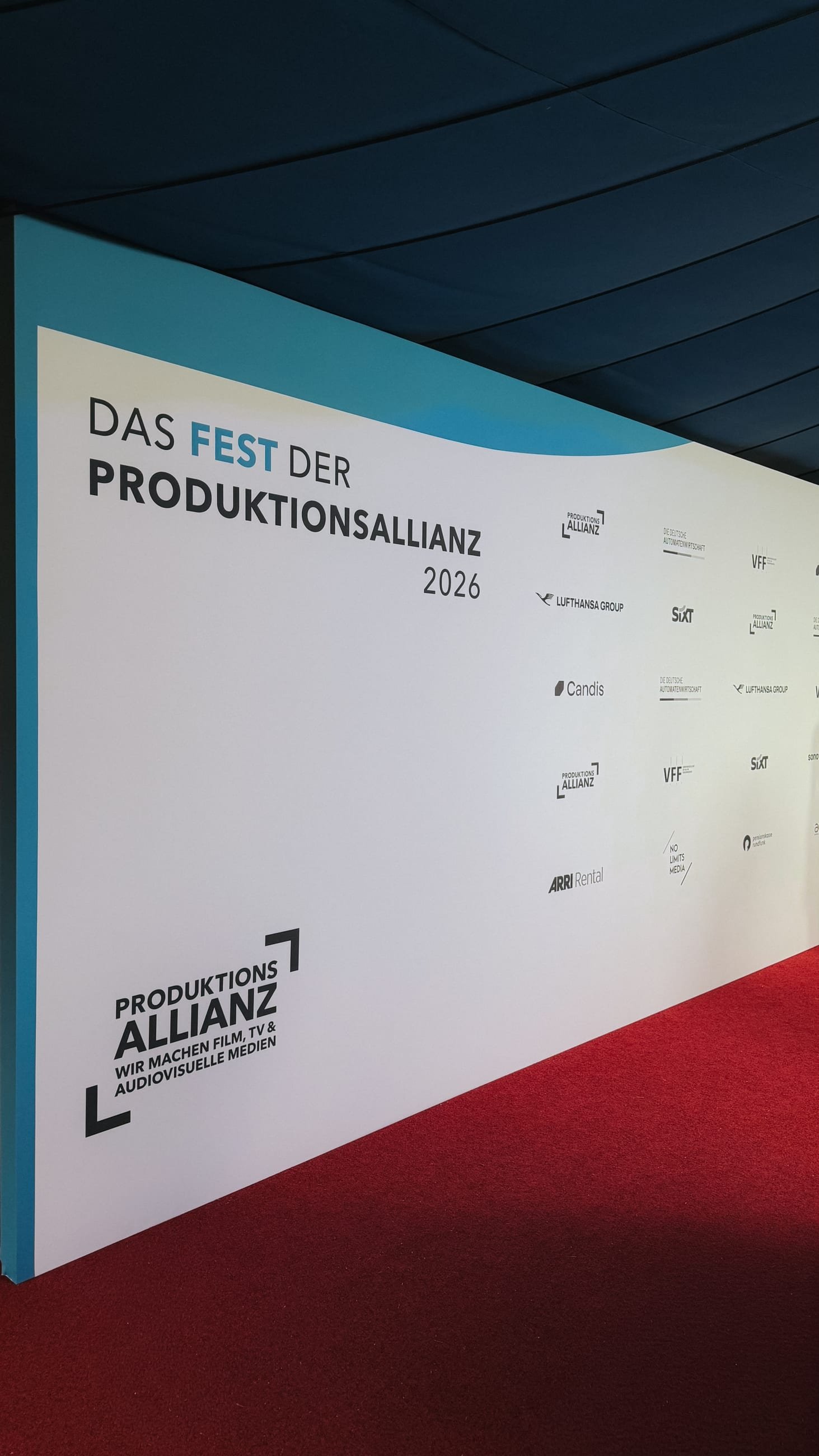 Photo wall reading „Das Fest der Produktionsallianz 2026“ with the partner logos, red carpet in front of it and the blue tent ceiling above