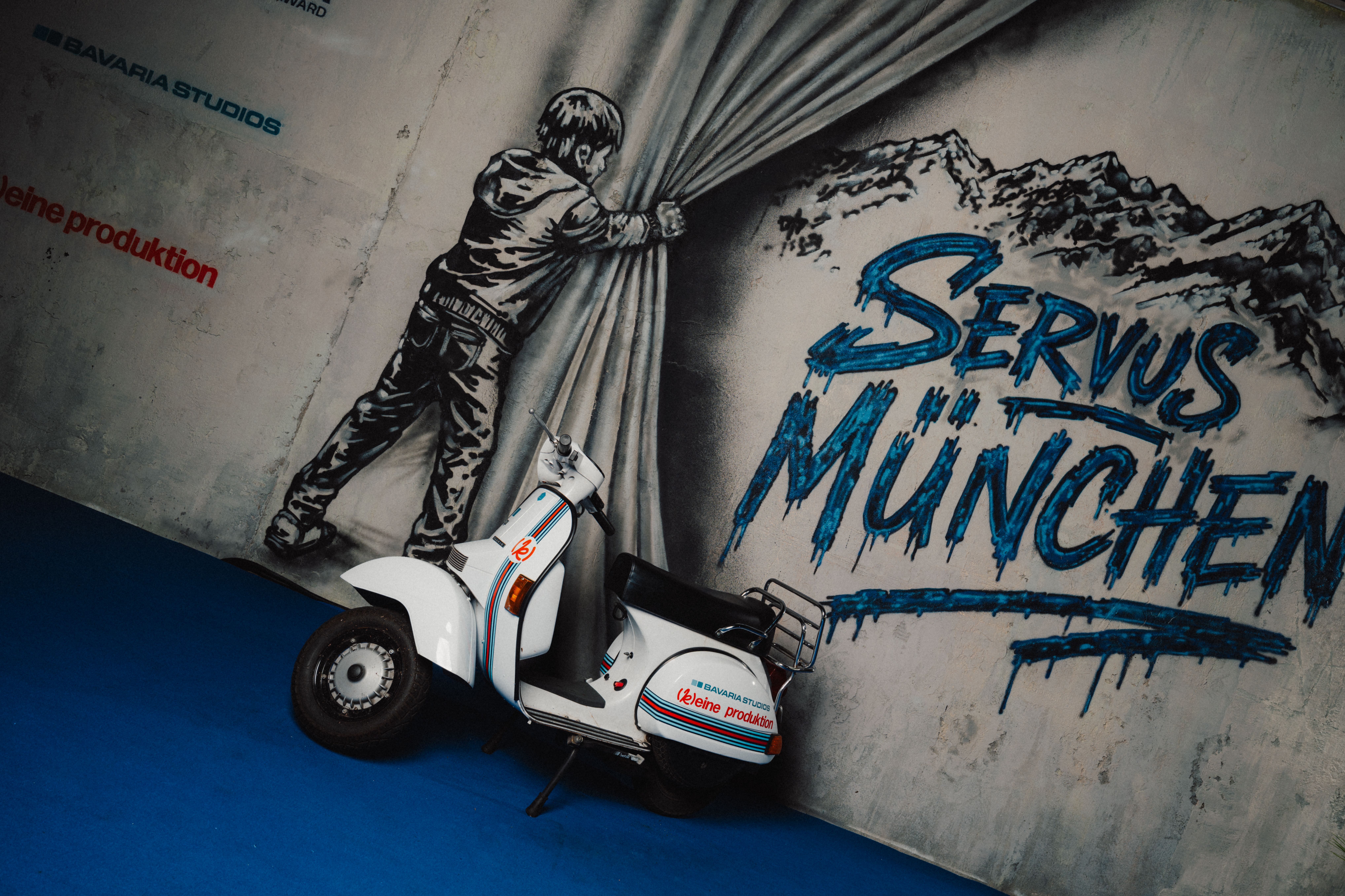 The branded Vespa of [k]eine produktion in front of the Servus München mural at the Bavaria Studios