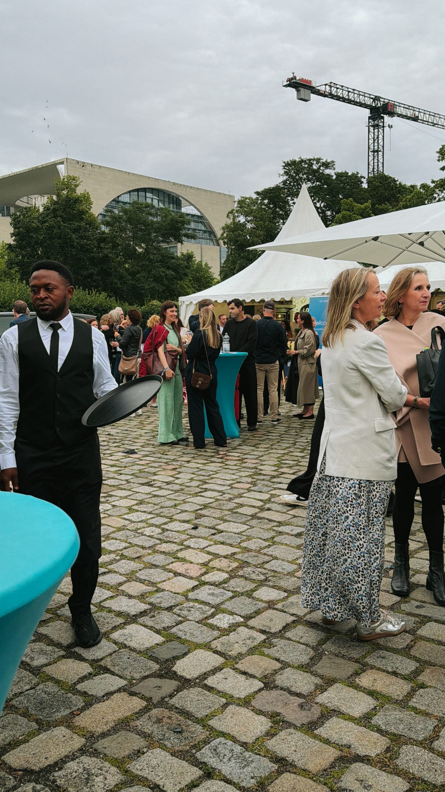 Guests and a waiter with a tray in the courtyard, white pagoda tents, the Federal Chancellery and a construction crane behind them