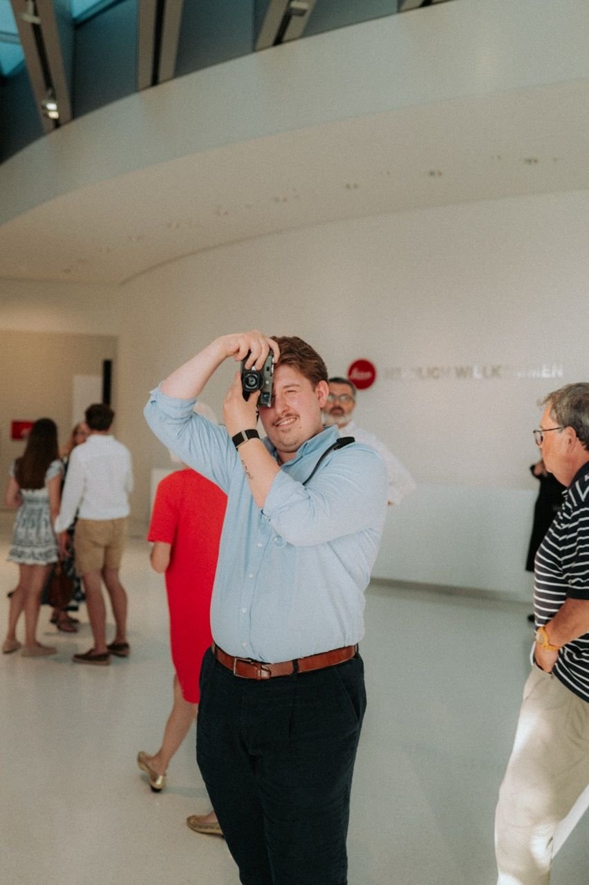 Person raising a Leica camera to their eye, taking a photo along the gallery walkway