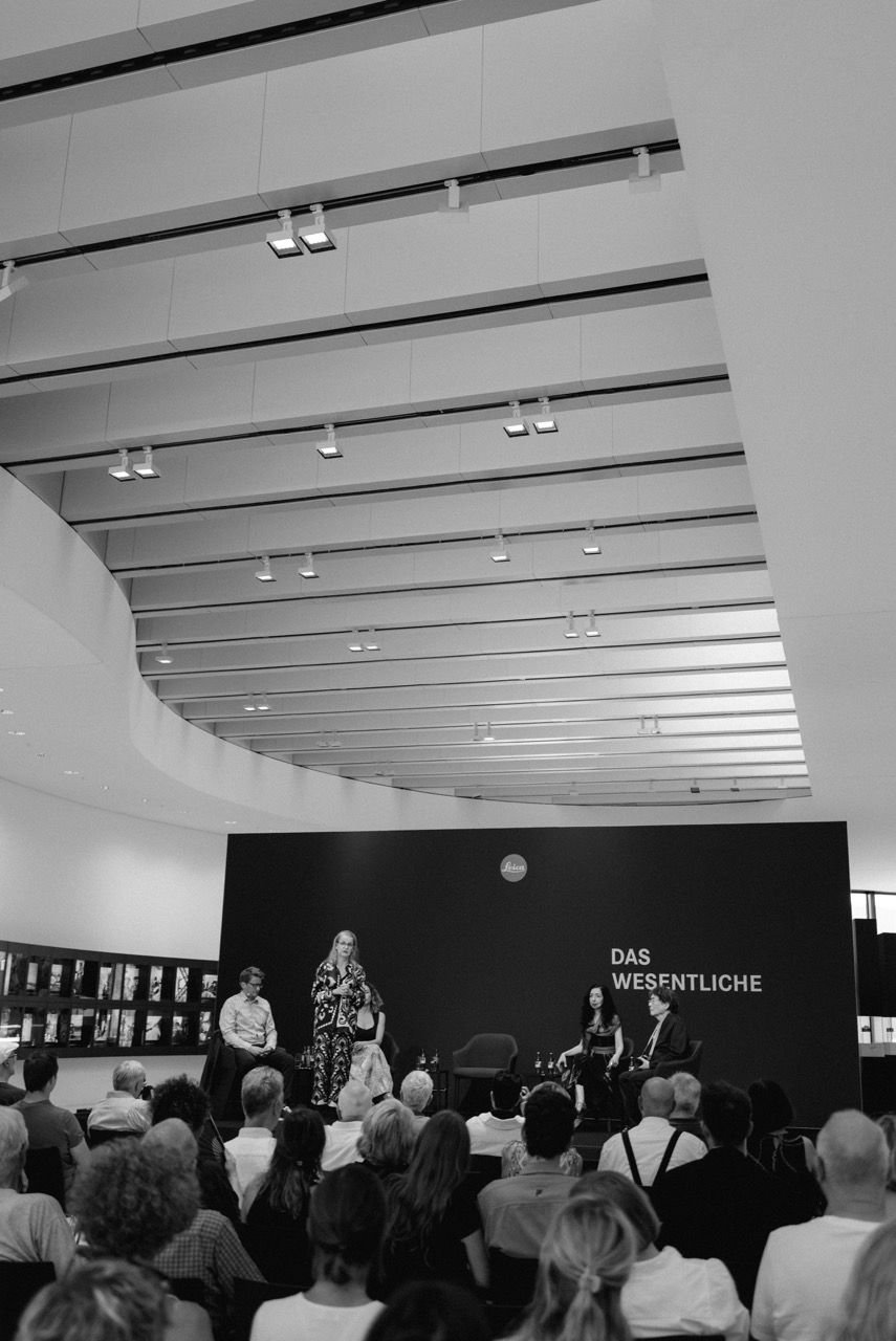 Panel of four people in front of a full audience, behind them a dark wall with the Leica logo and the words „Das Wesentliche“