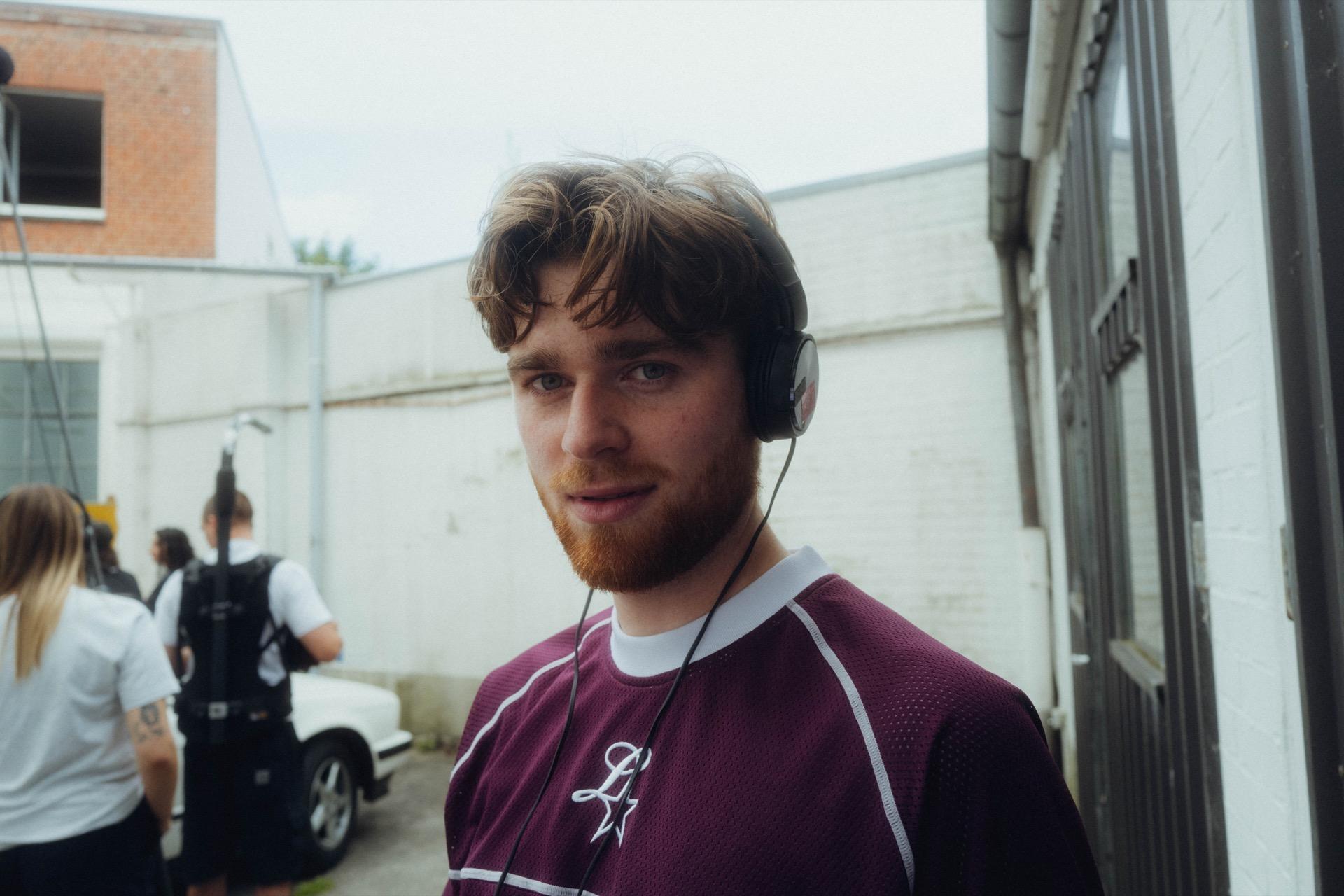 Portrait: a young man with a reddish beard and headphones in the burgundy Elstar jersey looks into the camera, the set blurred behind him