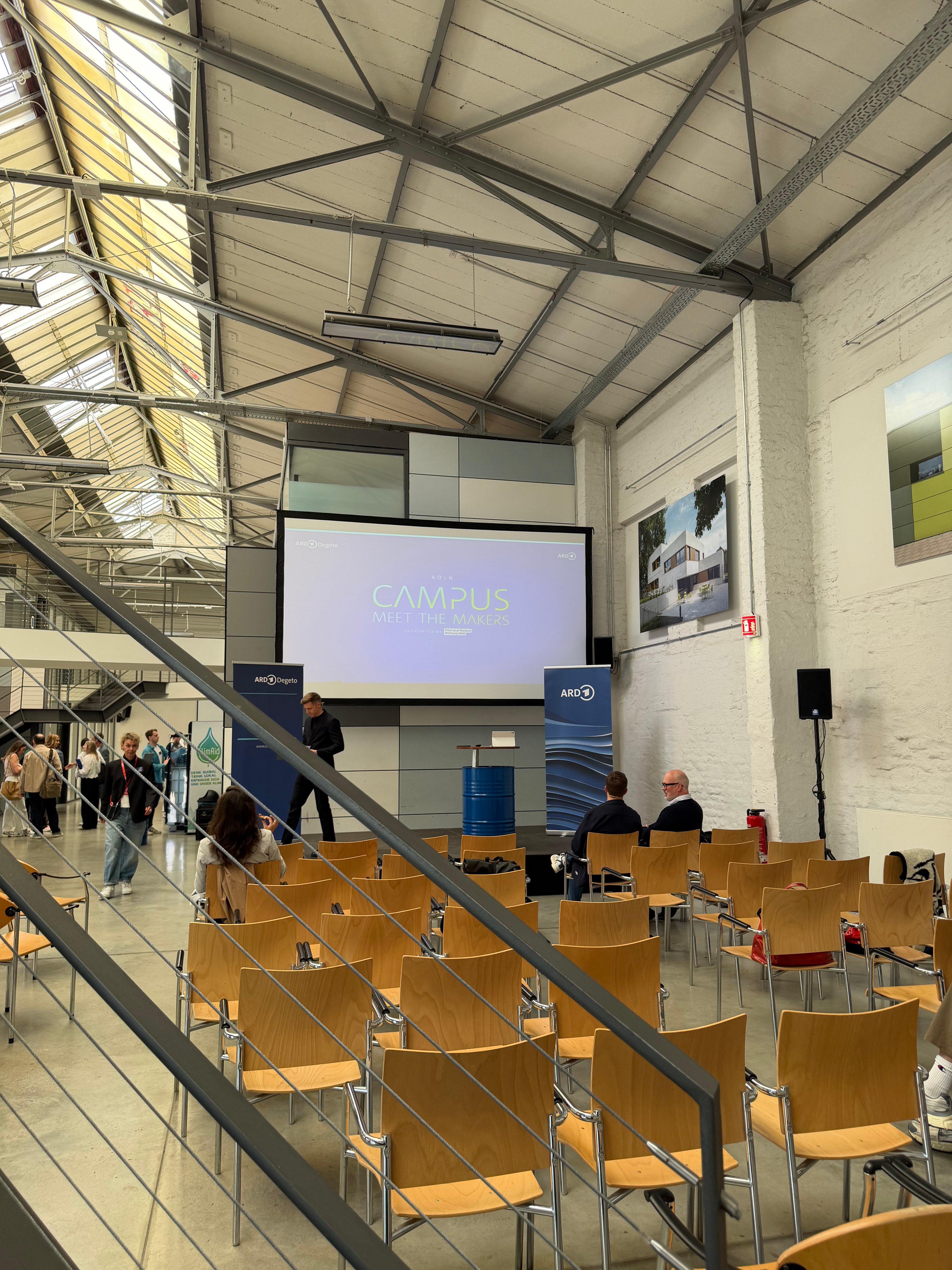 Blick in die Halle mit Stuhlreihen, Leinwand und ersten Gästen vor Beginn