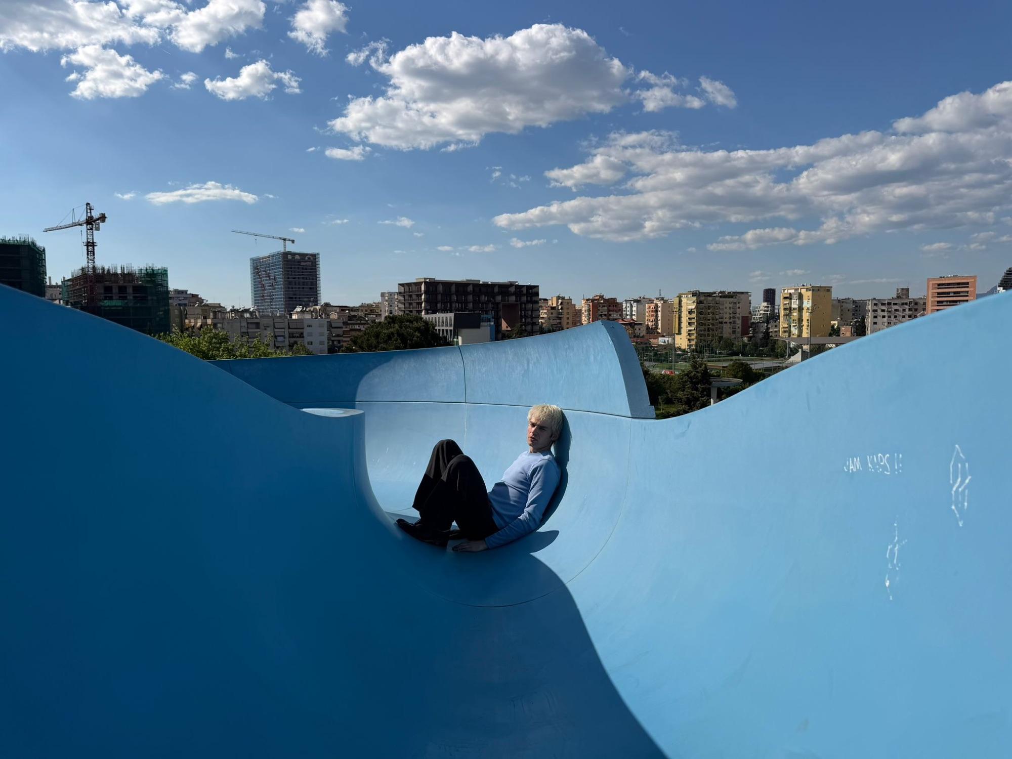 Jaschu sitzt in einem leeren blauen Betonbecken, dahinter die Skyline von Tirana mit Baukränen