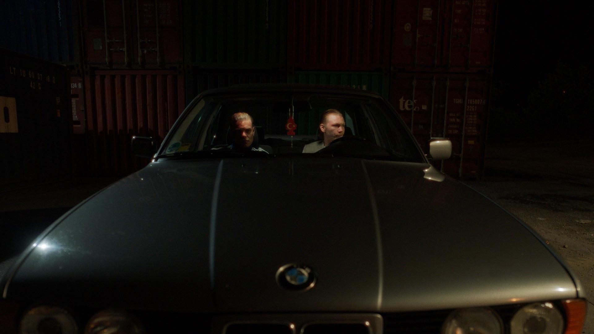 Two men sit at night in an old BMW in front of a wall of freight containers