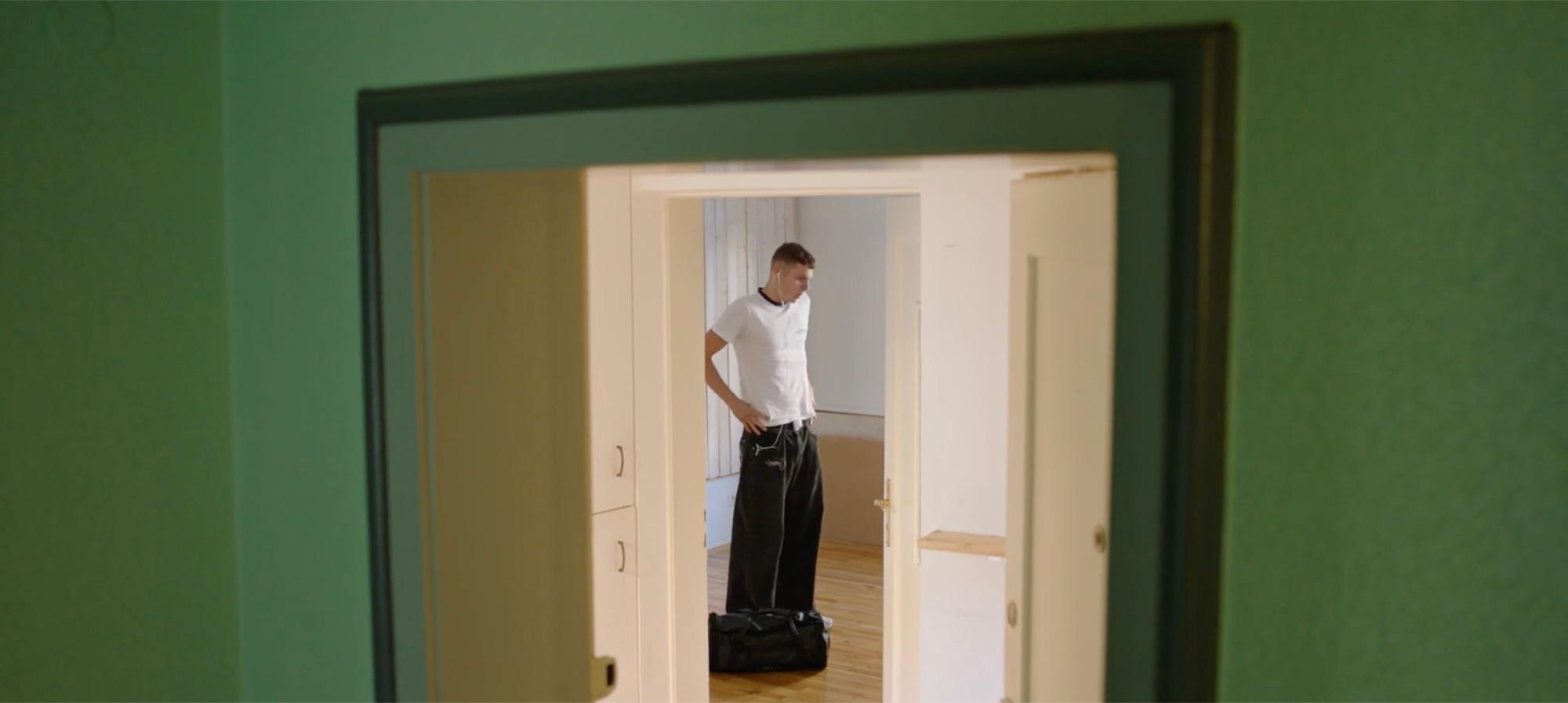 Moritz blickt zu Boden, allein im leeren Zimmer neben seiner Tasche, durch die Türöffnungen gerahmt