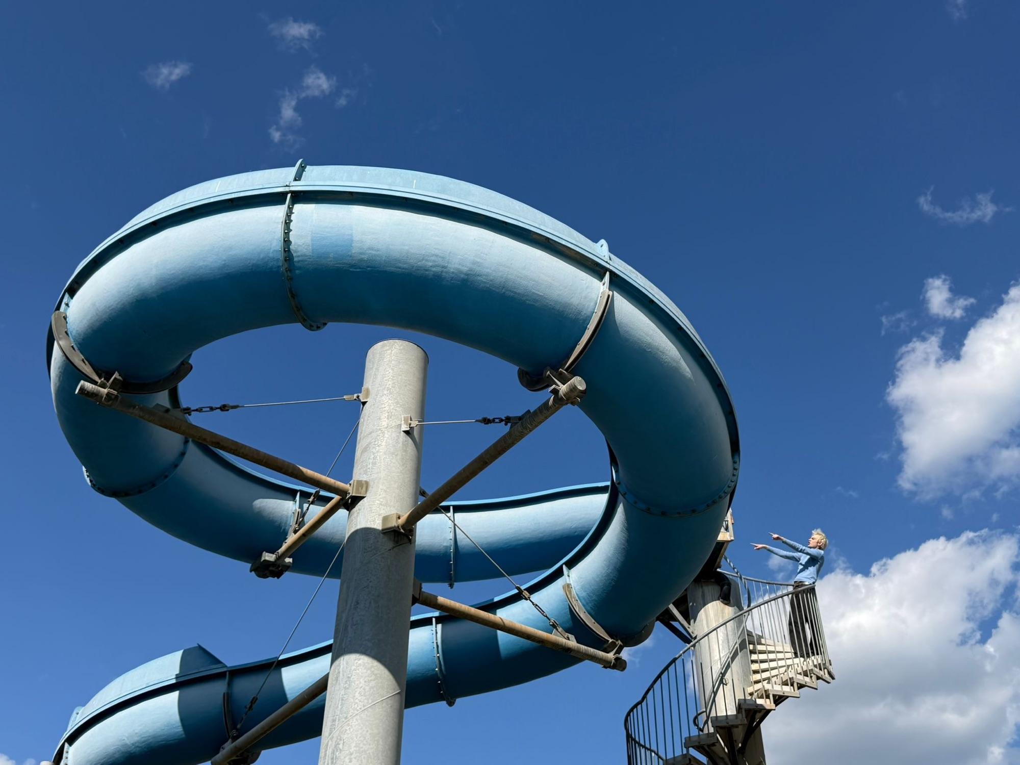 Eine blaue Wasserrutsche windet sich gegen den Himmel, oben steht Jaschu winzig auf der Treppe