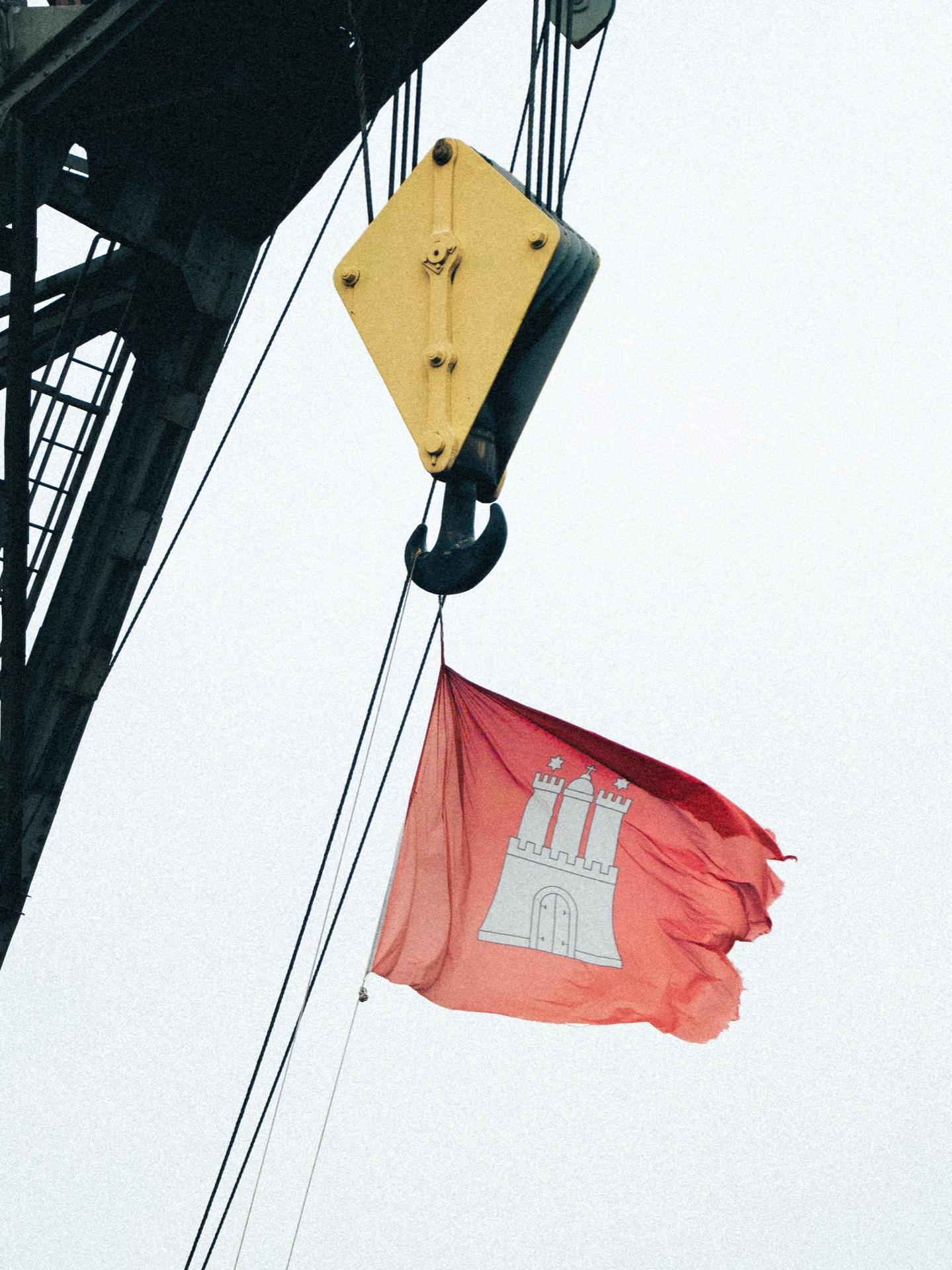 Detail: die rot-weiße Hamburger Flagge mit Burg-Wappen weht an einem Kranhaken vor weißem Himmel