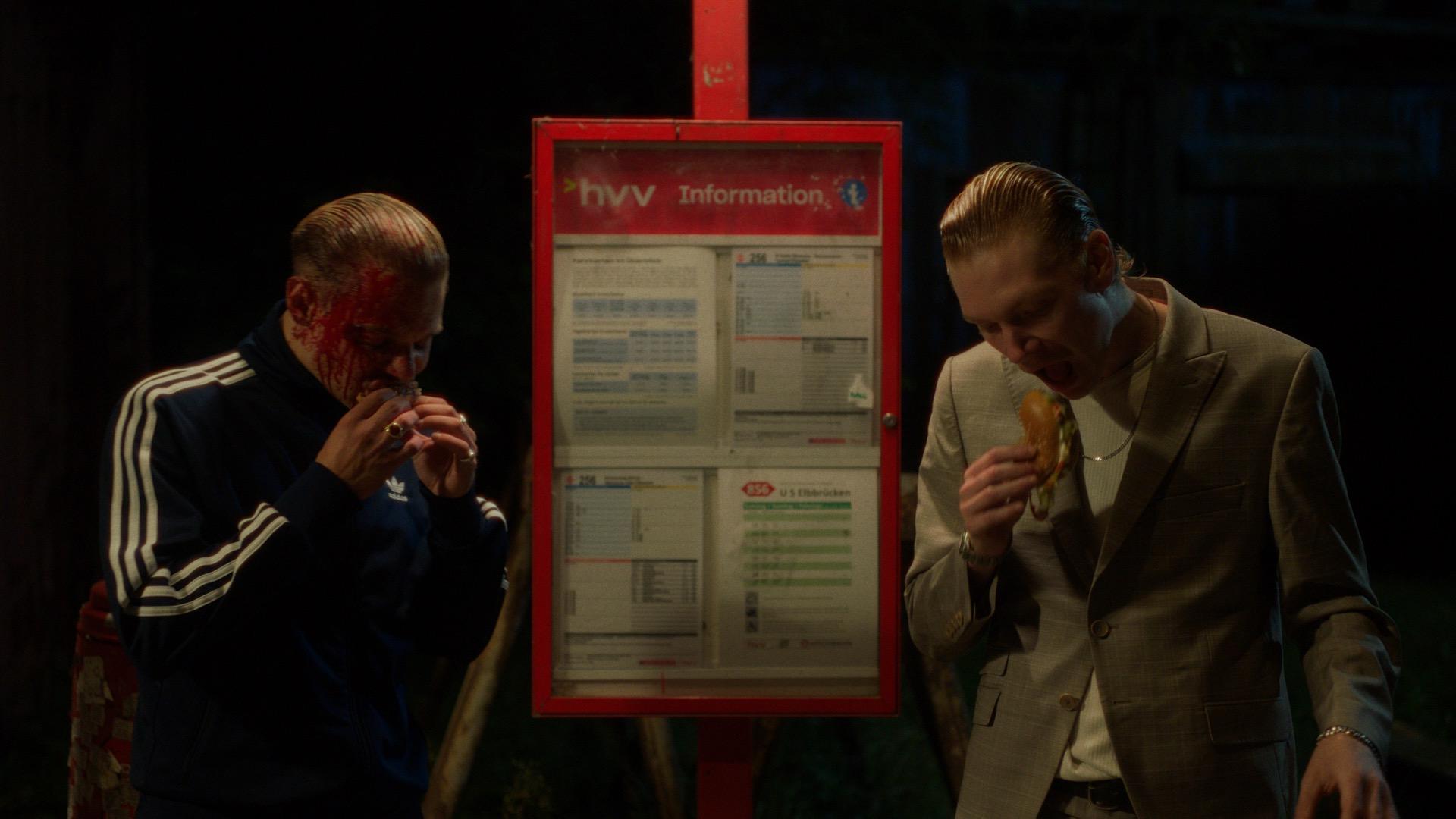 Two men eat at a HVV stop at night, one with blood smeared across his face