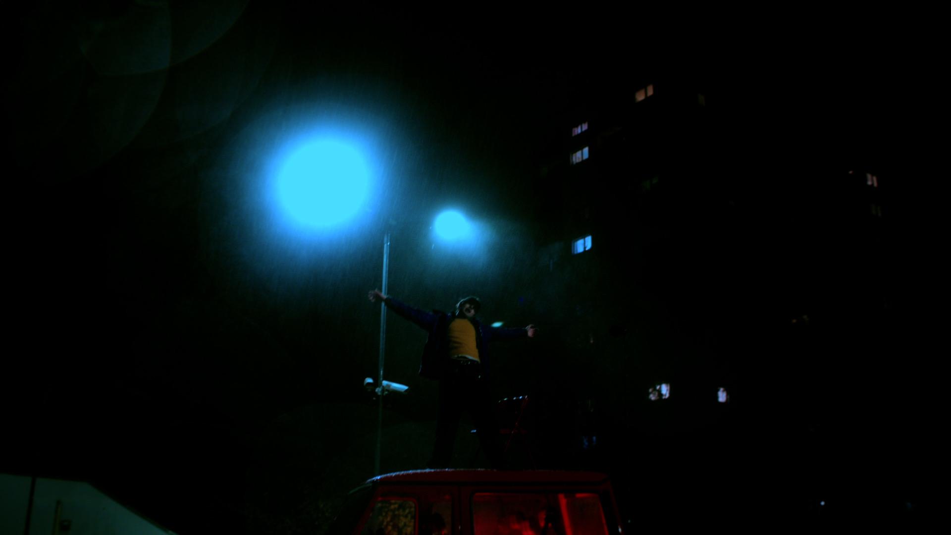 Jaschu stands with arms outstretched on the roof under two blue lights