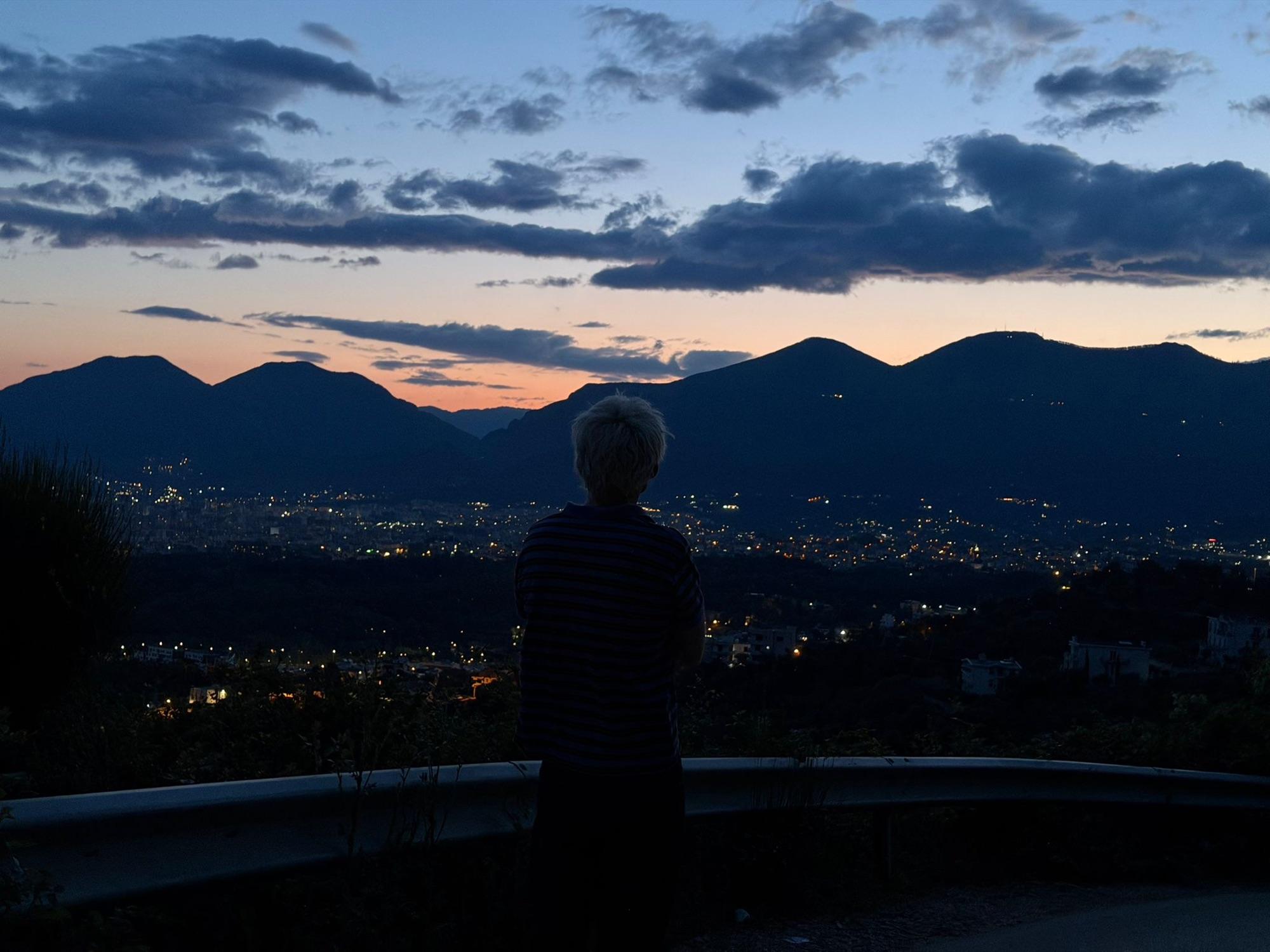 Jaschu als Silhouette an einer Leitplanke, davor eine Stadt und Berge im Sonnenuntergang