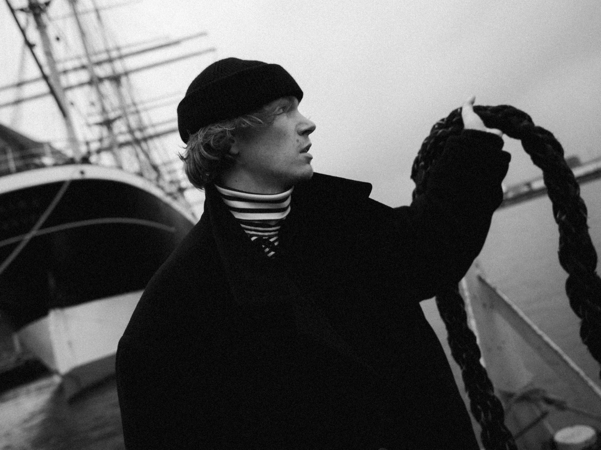 Schwarzweiß: Filip Schnack im Profil mit Schiffermütze an Deck eines Segelschiffs, Takelage und Hafen dahinter
