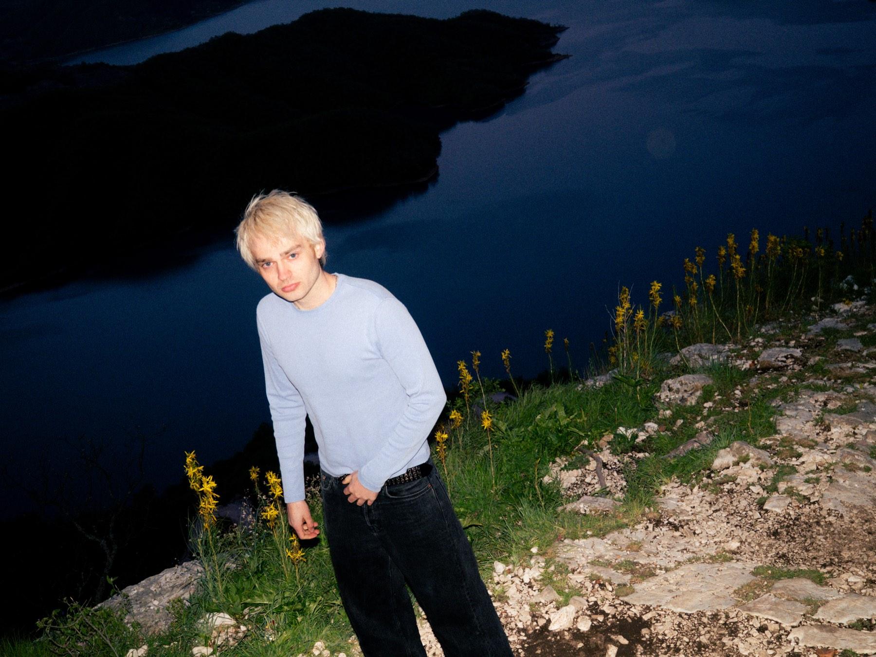Jaschu am Ufer eines dunklen Sees in der Nacht, hinter ihm das Wasser und die Hügel