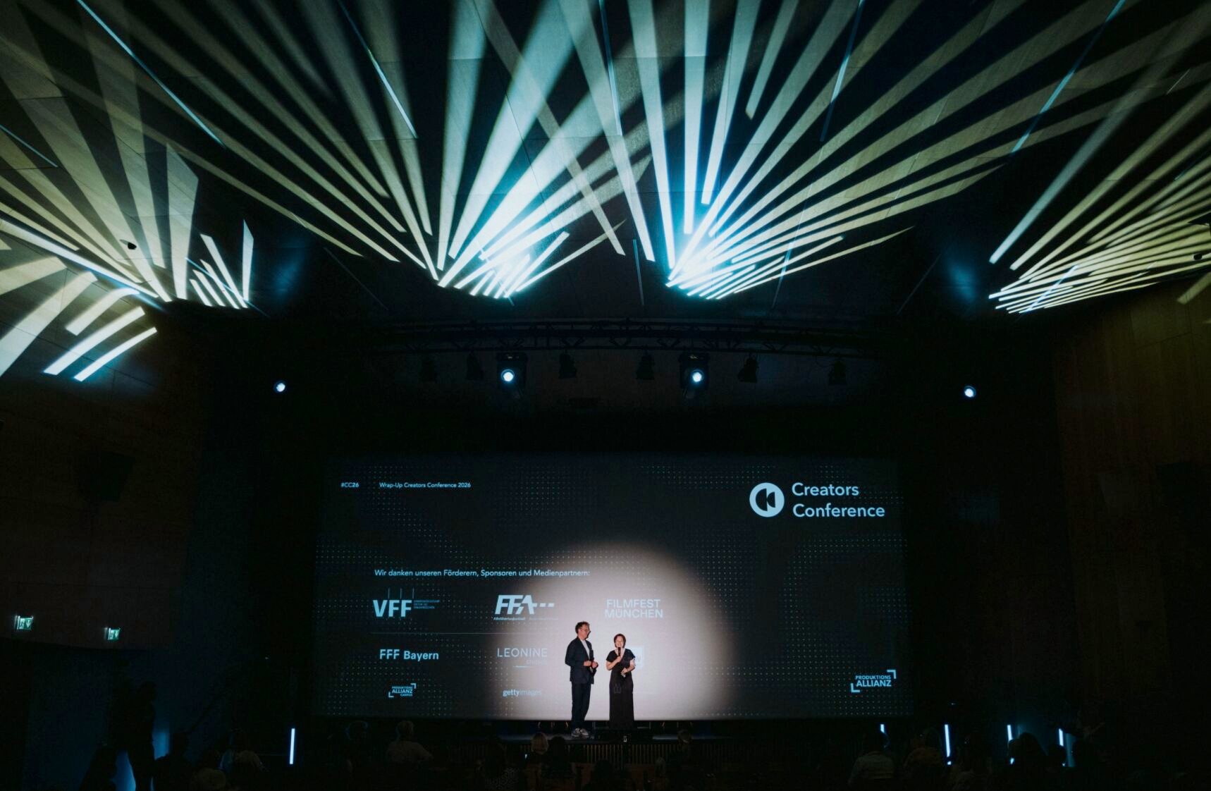 Bühne beim Wrap-Up der Creators Conference, über dem Saal ein Lichtdesign aus hellen Strahlen, auf der Leinwand die Logos der Förderer und Partner